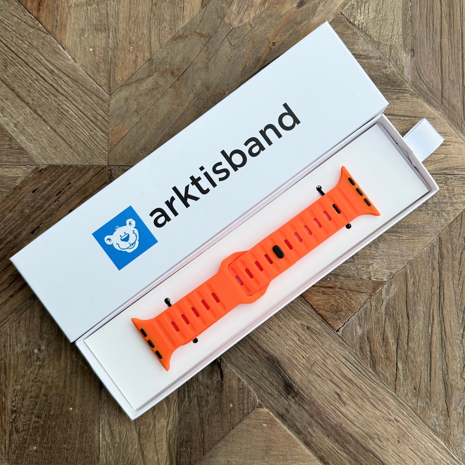 Ein orangefarbenes Arktisband Apple Watch Silikonarmband „Active“ mit atmungsaktivem Design wird in einer weißen Arktisband-Verpackung mit einem blauen Bärenlogo gezeigt, das auf einer hölzernen Oberfläche mit Fischgrätenmuster ruht.