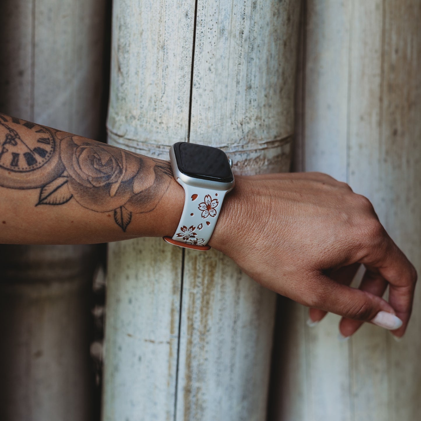 Der linke Arm einer Person mit einem Blumentattoo und einem Arktisband-Apple-Watch-Armband „Flores“ mit einem weißen Band mit kleinen roten und schwarzen Blumen, das vor vertikalen Bambusstäben gehalten wird.