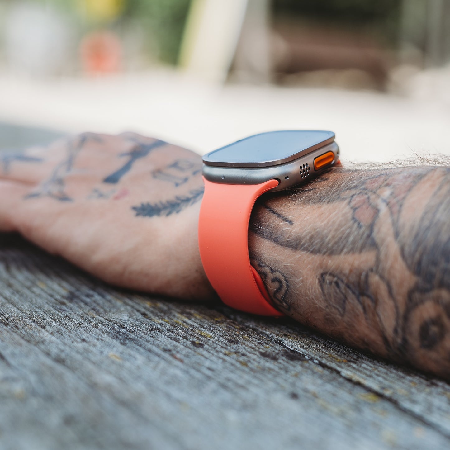 Eine Nahaufnahme eines tätowierten Arms, der auf Holz ruht und das arktisband Apple Watch Silikonarmband "Aqua" von arktisband trägt. Das Display ist ausgeschaltet und der Hintergrund unscharf.