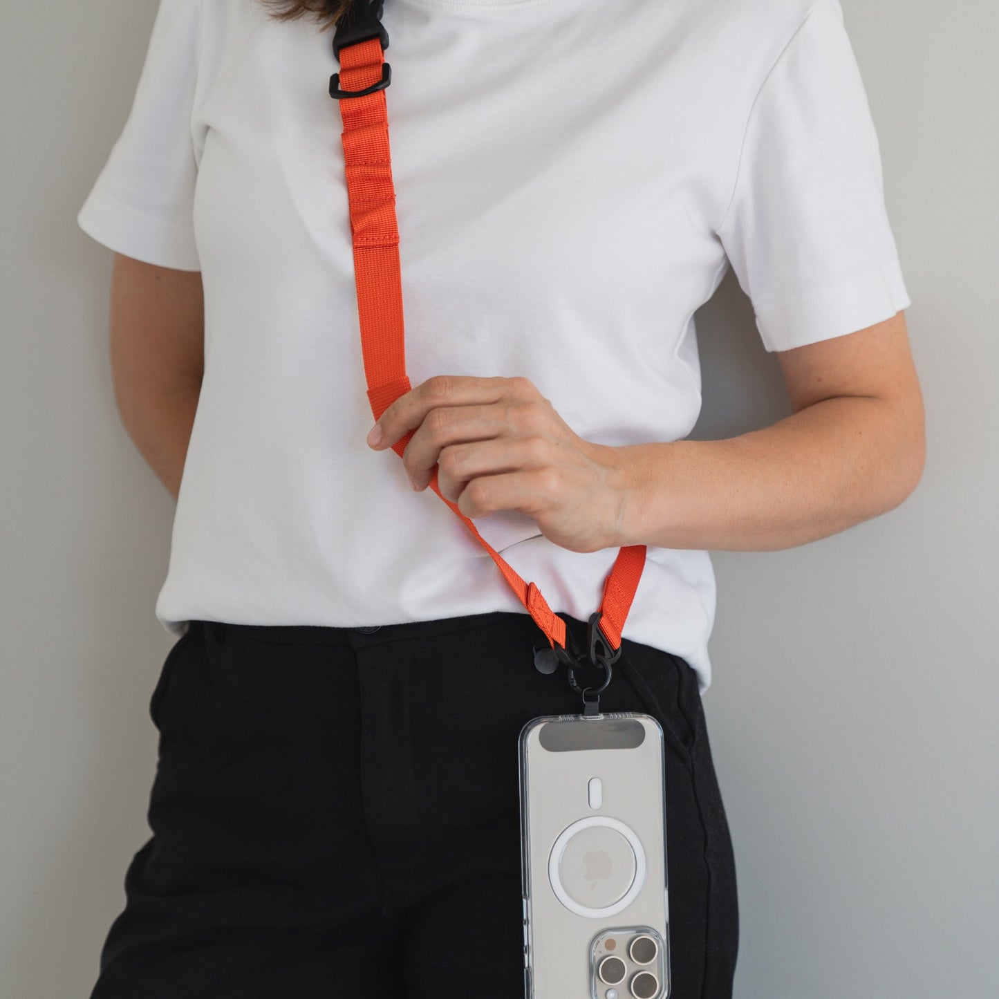 Eine Person in weißem T-Shirt und schwarzer Hose trägt ein arktis Smartphone-Gurtband-Lanyard, befestigt an einer transparenten Handyhülle mit 1-Klick-Verschluss im Crossbody-Stil. Der Hintergrund ist eine schlichte, helle Wand.