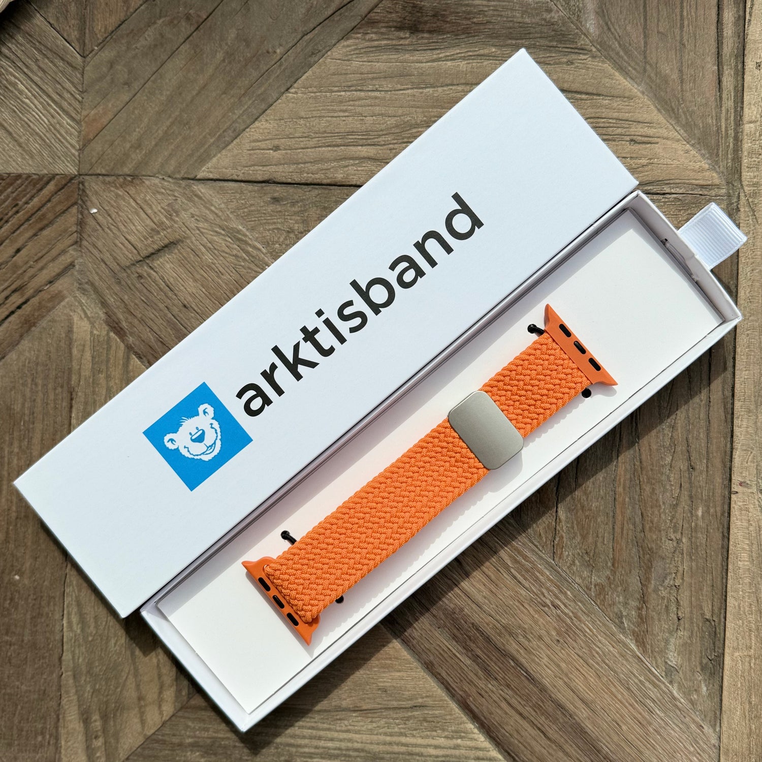 Ein von arktisband geflochtenes Apple Watch Armband „Casual“ mit Magnetverschluss in Orange wird in seiner arktisband-Box auf einer Holzoberfläche präsentiert.