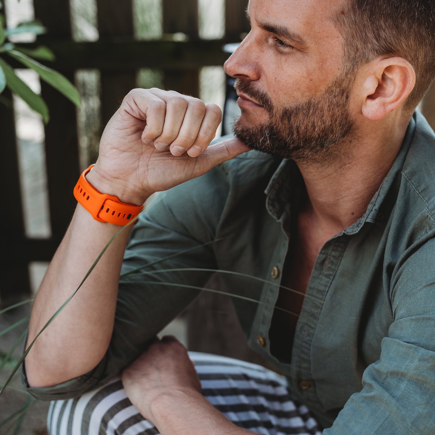 Ein Mann mit kurzen Haaren und Bart sitzt im Freien. Er trägt ein grünes Hemd, eine gestreifte Hose und ein arktisband Apple Watch FKM Sportarmband "Sweat" von arktisband. Im Hintergrund sind ein Holzzaun und Pflanzen zu sehen.