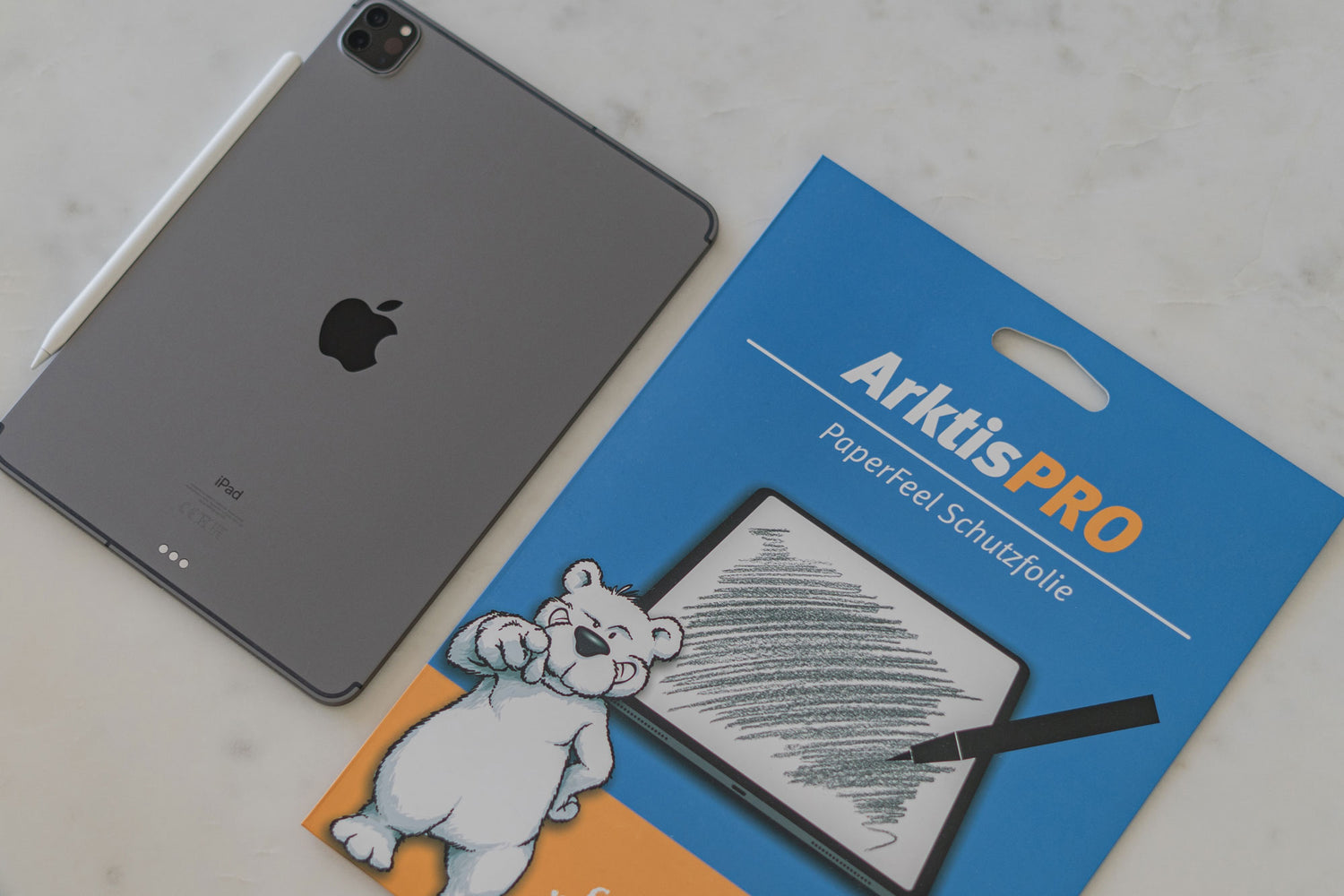 Ein graues iPad mit einem weißen Stift neben der Verpackung des ArktisPRO Paperfeel-Displayschutzes auf einem Tisch.