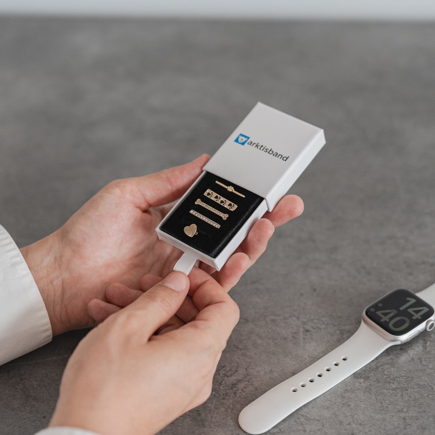 Eine Person öffnet eine kleine weiße Schachtel mit der Aufschrift „okHusband“. Darin befinden sich goldene Arktisband Apple Watch Charms „Pet Love“. Eine weiße Smartwatch, ideal für die Kombination mit diesen Arktisband-Accessoires, liegt daneben auf der grauen Fläche.
