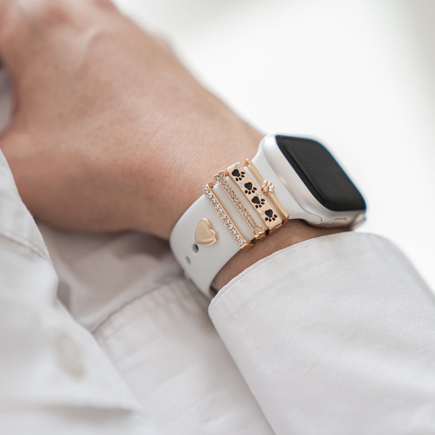 Eine Person trägt das arktisband Apple Watch Armband Charms mit goldenen und silbernen Anhängern wie Herz, Pfote und Edelsteinen. Das exklusive Künstler-Accessoire rundet den Look zum weißen Langarmshirt individuell ab.