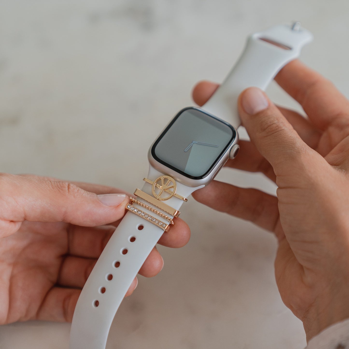 Eine Person hält eine Apple Watch mit einem weißen Arktisband-Armband mit goldenen Arktisband-„Peace“-Anhängern, die mit dekorativen goldenen und diamantähnlichen Akzenten verziert sind. Das Zifferblatt ist bis auf einfache Uhrzeiger leer.