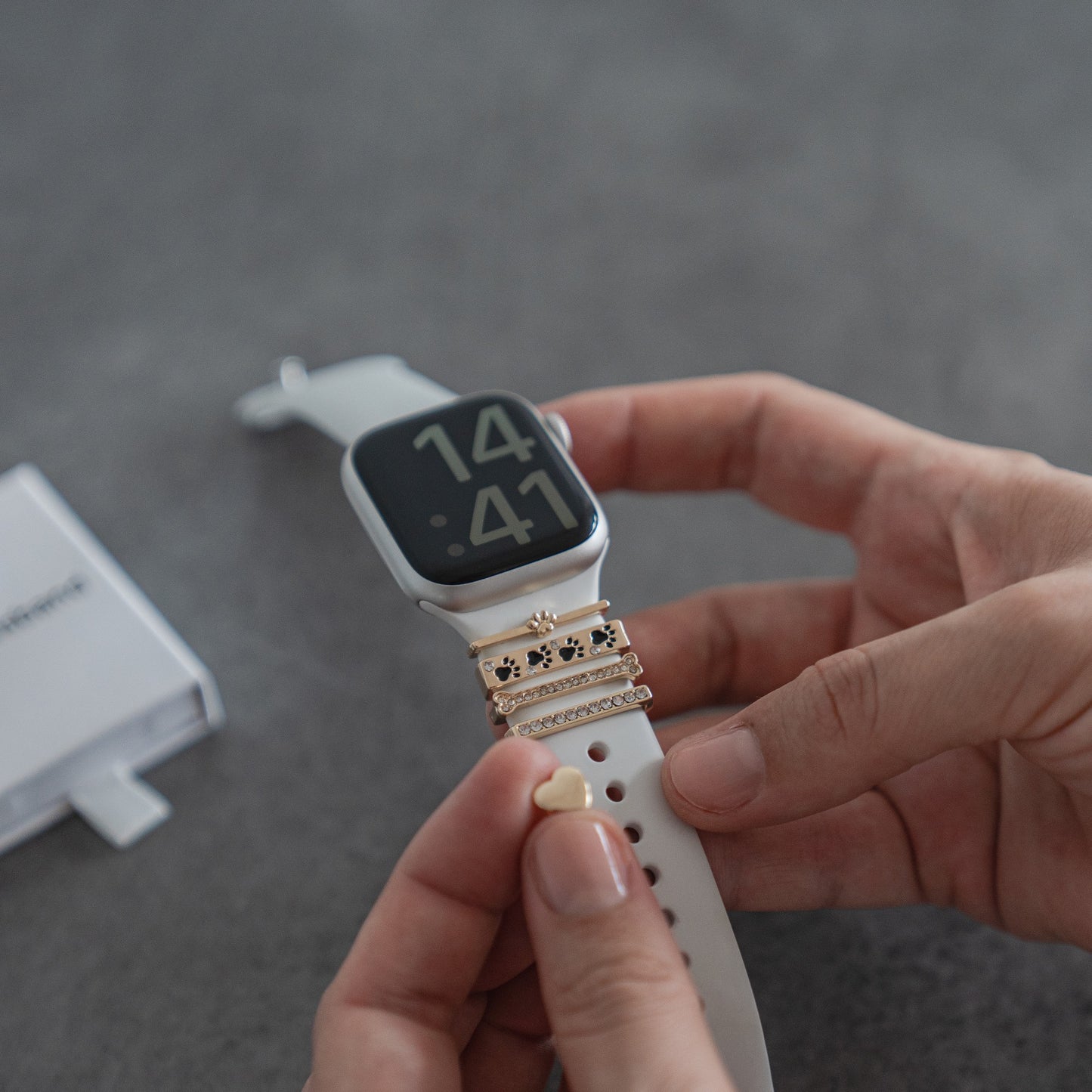 Eine Person befestigt die goldenen Arktisband Apple Watch Charms „Pet Love“ an einem weißen Arktisband ihrer Smartwatch, die bereits über mehrere dekorative Apple Watch-Accessoires verfügt, auf einer grauen Oberfläche.