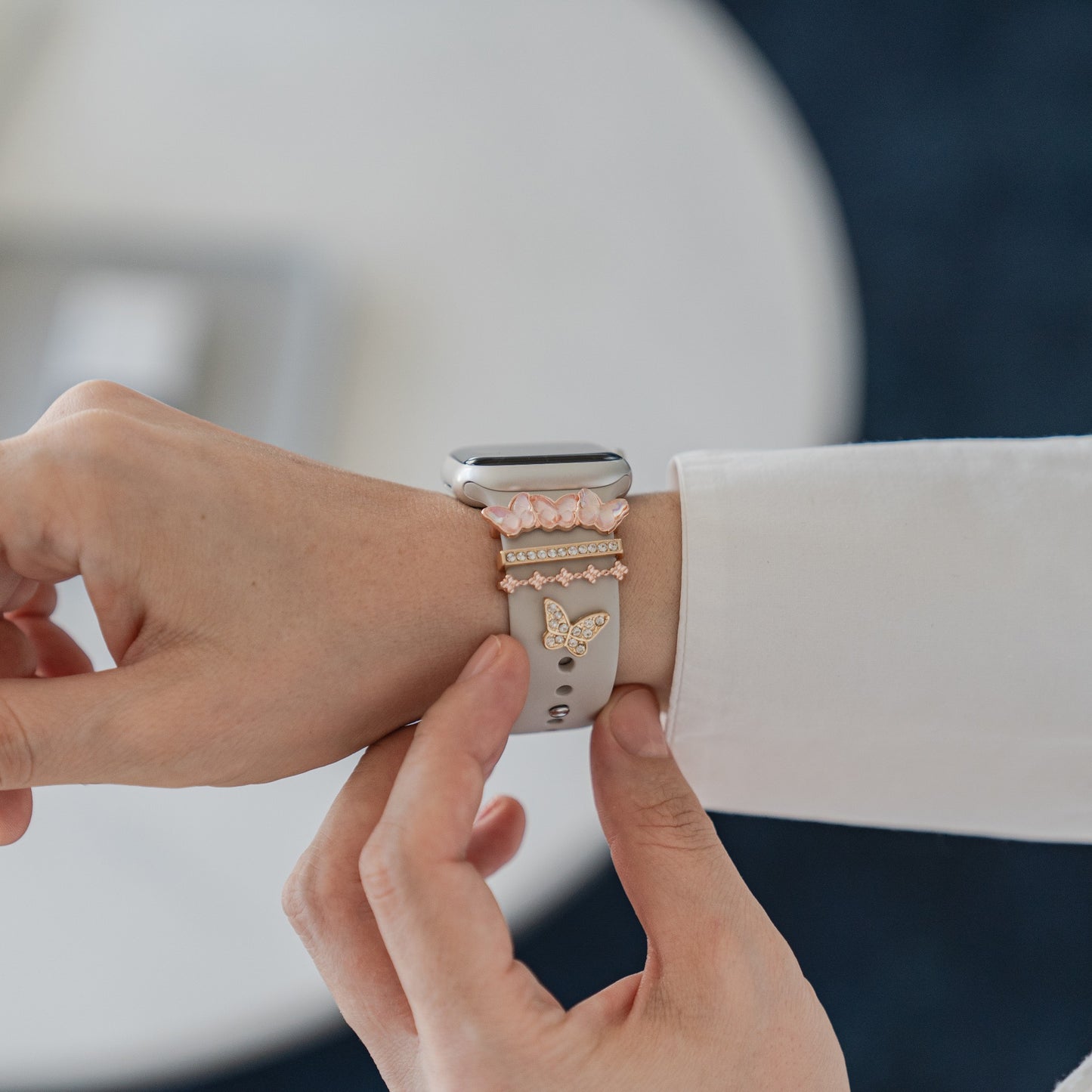 Eine Person befestigt ein Arktisband Apple Watch Armband Charms mit einem weißen, anpassbaren Armband und roségoldenen Schmetterlings- und Kristall-Charms von Arktisband an ihrem Handgelenk, mit einem unscharfen Hintergrund.