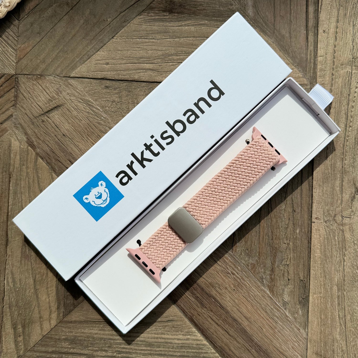 Das arktisband Geflochtene Apple Watch Armband „Casual“ mit Magnetverschluss in Pink präsentiert sich elegant in seiner weißen arktisband Box auf einer Holzoberfläche.
