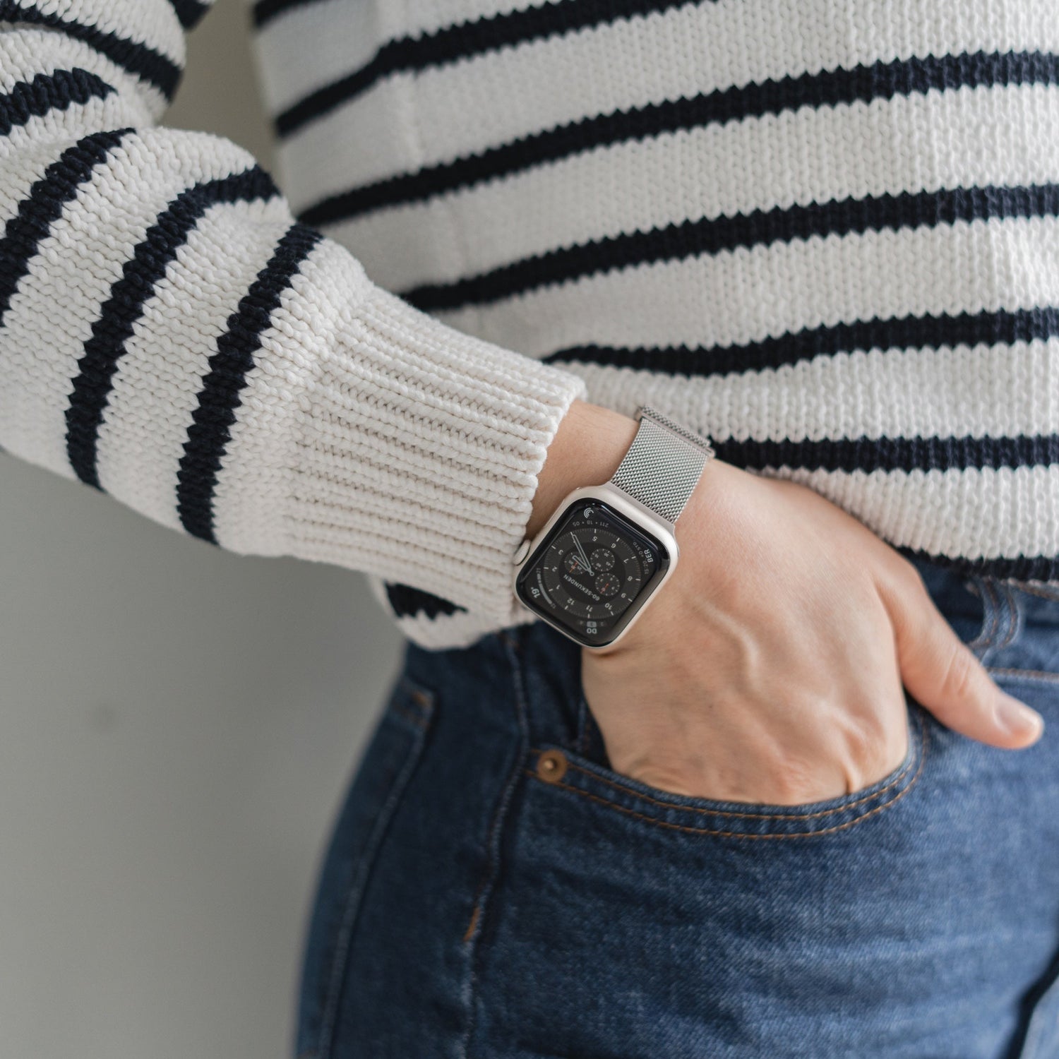 Eine Person in einem weiß-schwarz gestreiften Pullover und Bluejeans hat die Hand in der Tasche und präsentiert ein Arktisband Apple Watch Milanaise Loop Armband an ihrem Handgelenk.