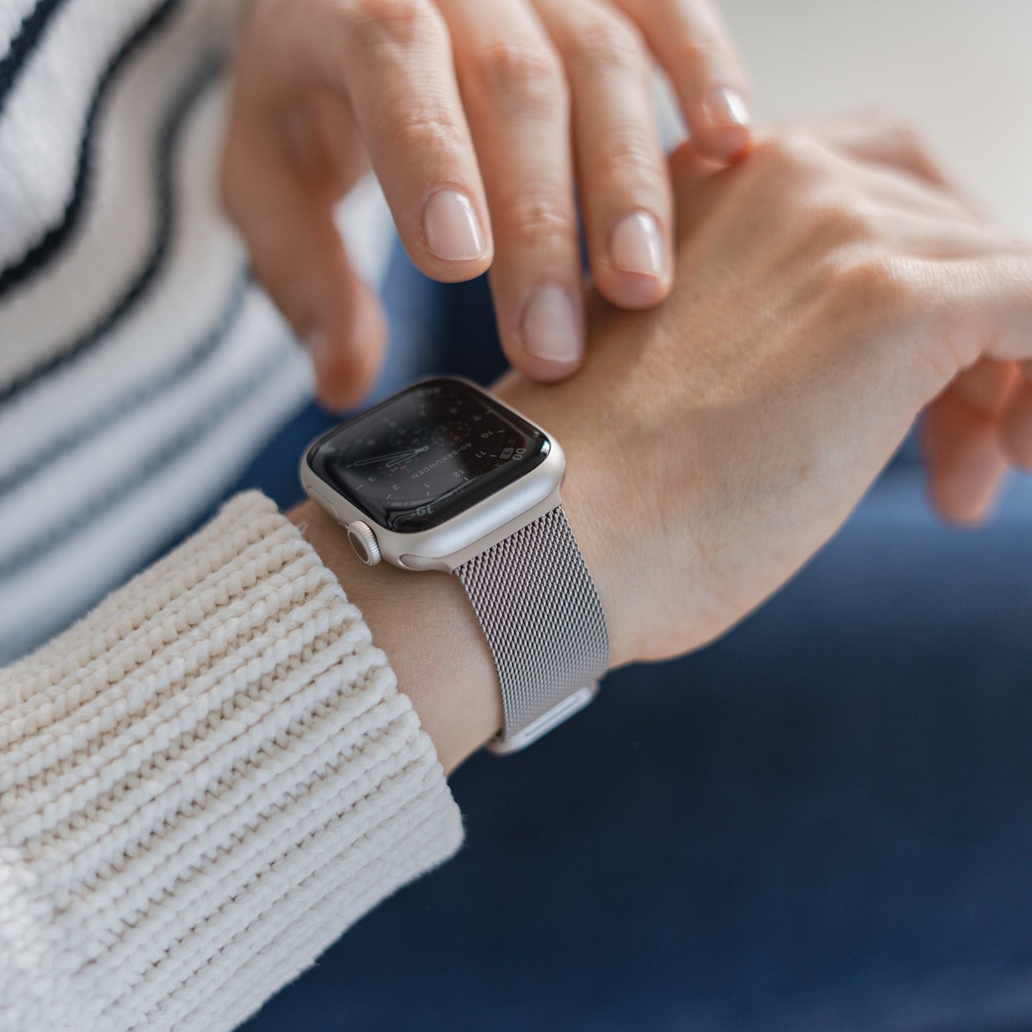 Eine Person in einem weißen Pullover mit schwarzen Streifen und blauen Hosen berührt mit der rechten Hand ihre Apple Watch, die mit einem arktisband Apple Watch Milanaise Loop Armband von arktisband gekoppelt ist.
