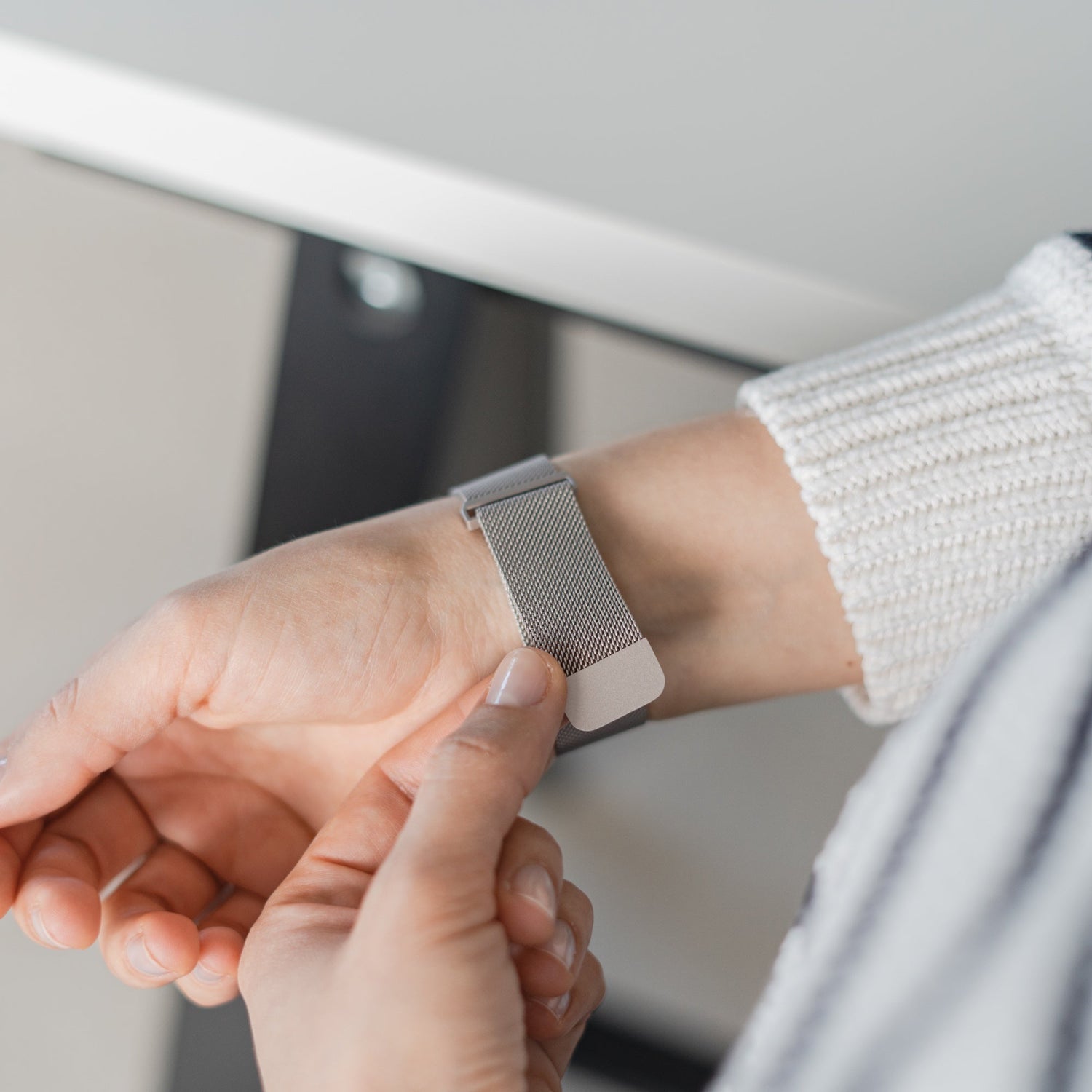 Eine Person befestigt ein Arktisband Apple Watch Milanaise Loop Armband an ihrem linken Handgelenk. Sie trägt einen weißen Strickpullover mit schwarzen Streifen.