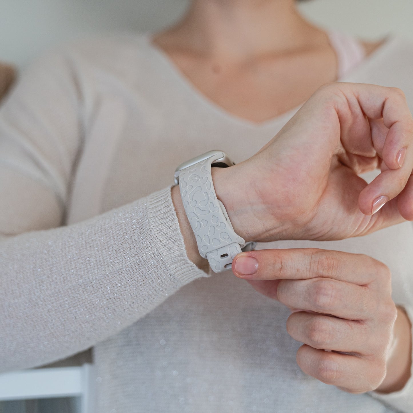 Eine Person befestigt das Arktisband Apple Watch Armband „Leopard“, ein hellgraues Silikonarmband mit dezentem Leopardenmuster, am linken Handgelenk und trägt einen cremefarbenen Langarmpullover. Der Fokus liegt auf den Zeigern und dem Apple Watch kompatiblen Armband.