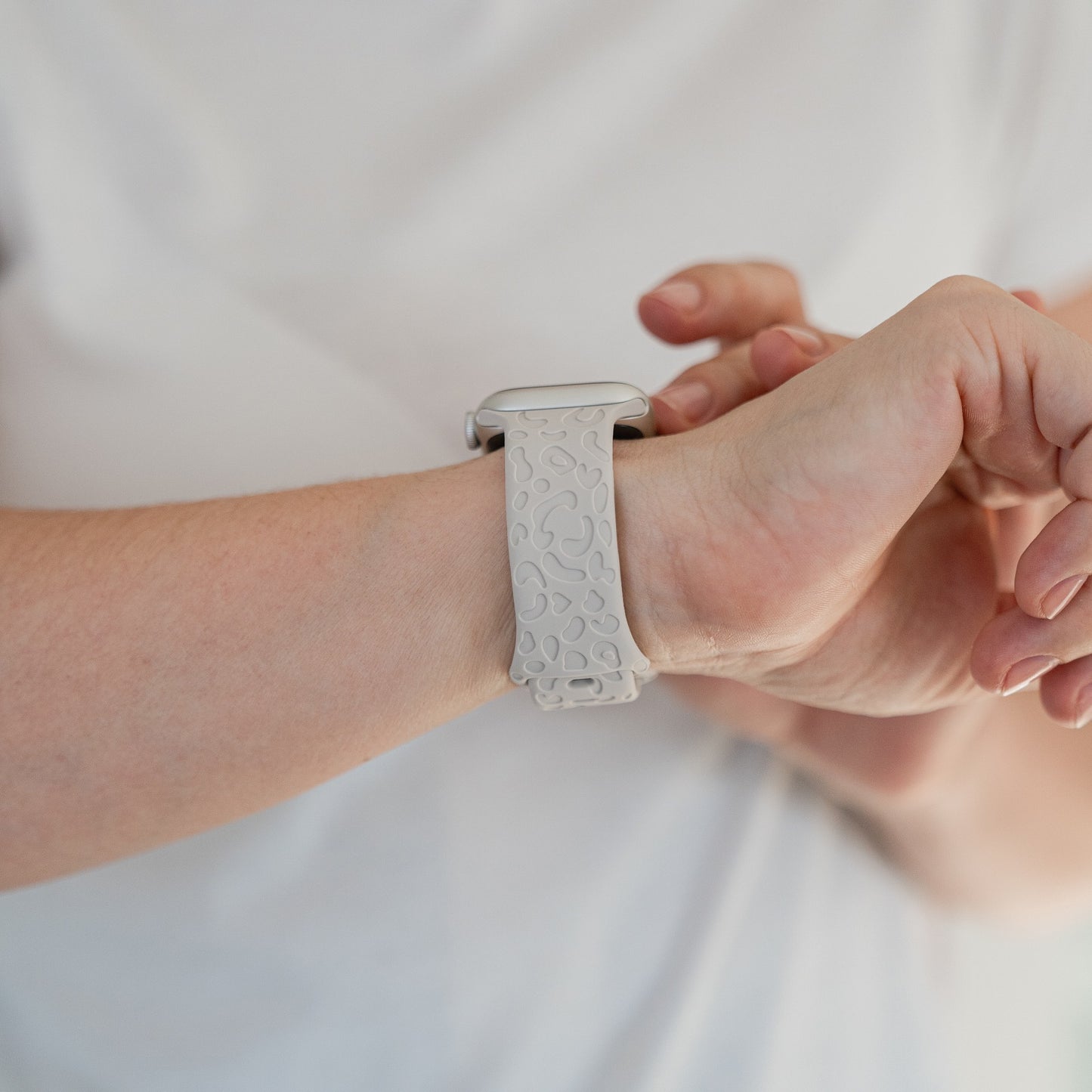 Eine Person im weißen Hemd rückt ihr arktisband Apple Watch Armband „Leopard“ von arktisband, ein hellgraues Silikonband mit erhabenem, abstraktem Muster, an ihrem linken Handgelenk zurecht.