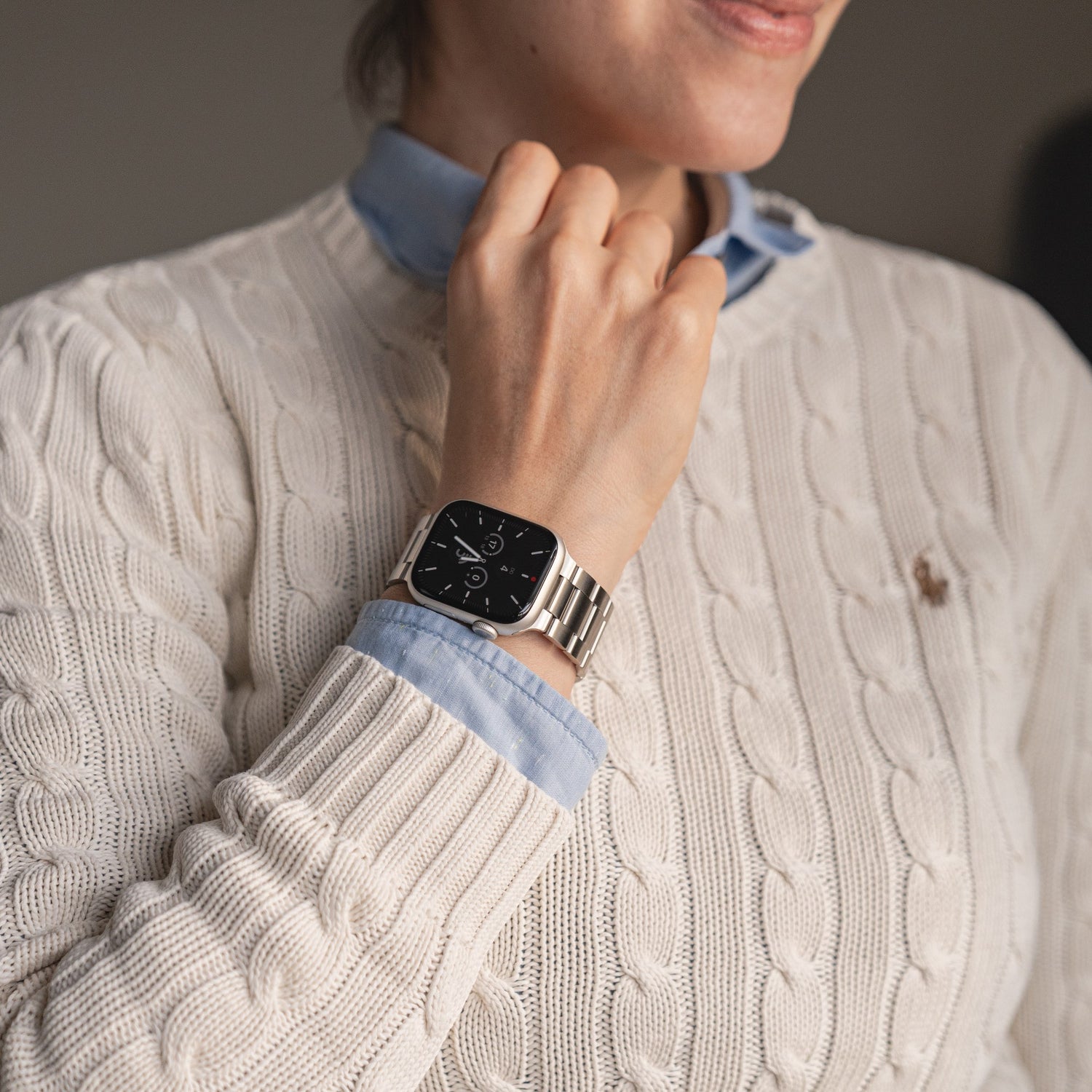 Eine Person in einem cremefarbenen Zopfmusterpullover und einem blauen Hemd präsentiert ihr Handgelenk. Sie trägt das arktisband Apple Watch Gliederarmband „Prime“ von arktisband mit einem analogen Zifferblatt auf dem schwarzen Bildschirm.