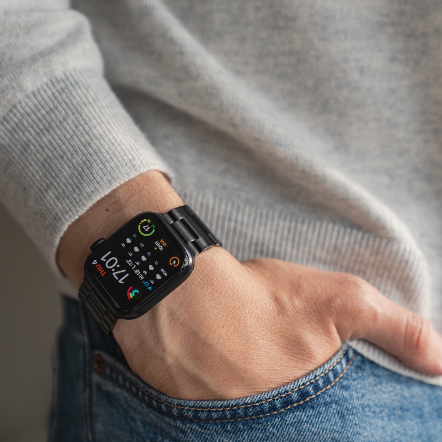 Eine Person in grauem Pullover und Bluejeans hat die linke Hand in der Hosentasche und präsentiert das arktisband Apple Watch Gliederarmband „Prime“ mit bunter Digitalanzeige am Handgelenk.