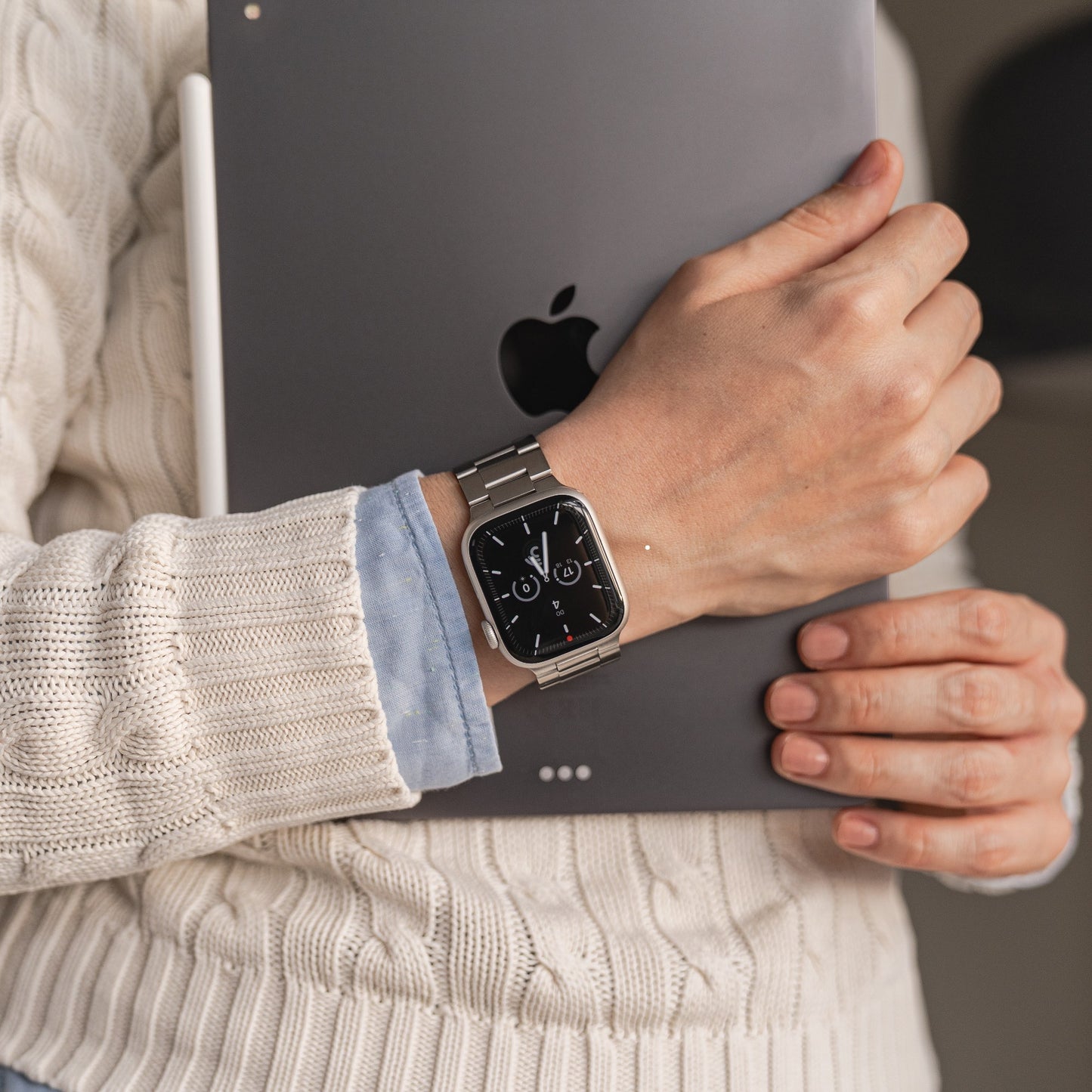 Eine Person in einem cremefarbenen Strickpullover trägt die Edelstahl-Smartwatch arktisband Apple Watch Gliederarmband „Prime“ von arktisband und hält ein geschlossenes iPad mit dem Apple-Logo nach außen.