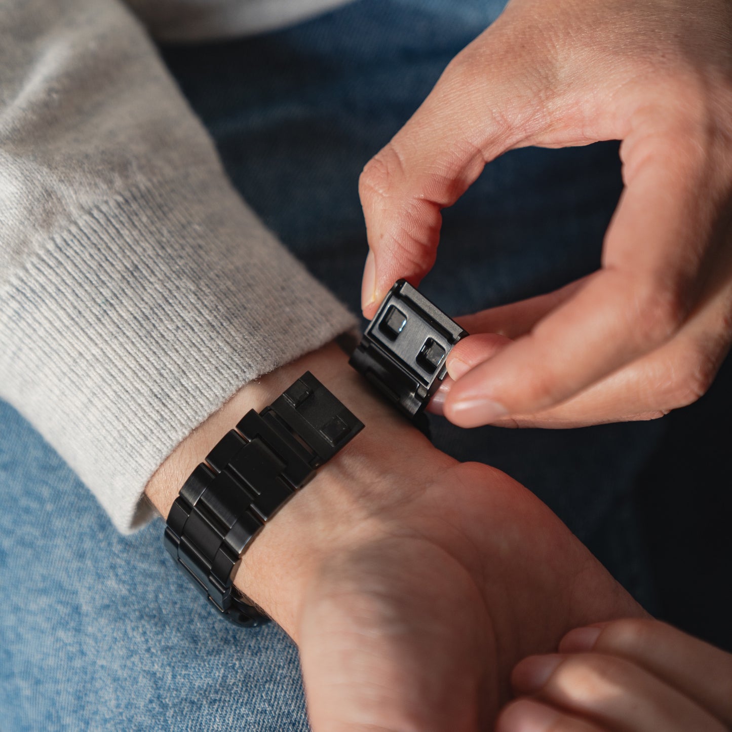 Eine Person befestigt das arktisband Apple Watch Gliederarmband „Prime“ mit Edelstahlarmband und MagneticClick Verschluss am Handgelenk und trägt ein hellgraues Langarmshirt und blaue Jeans.