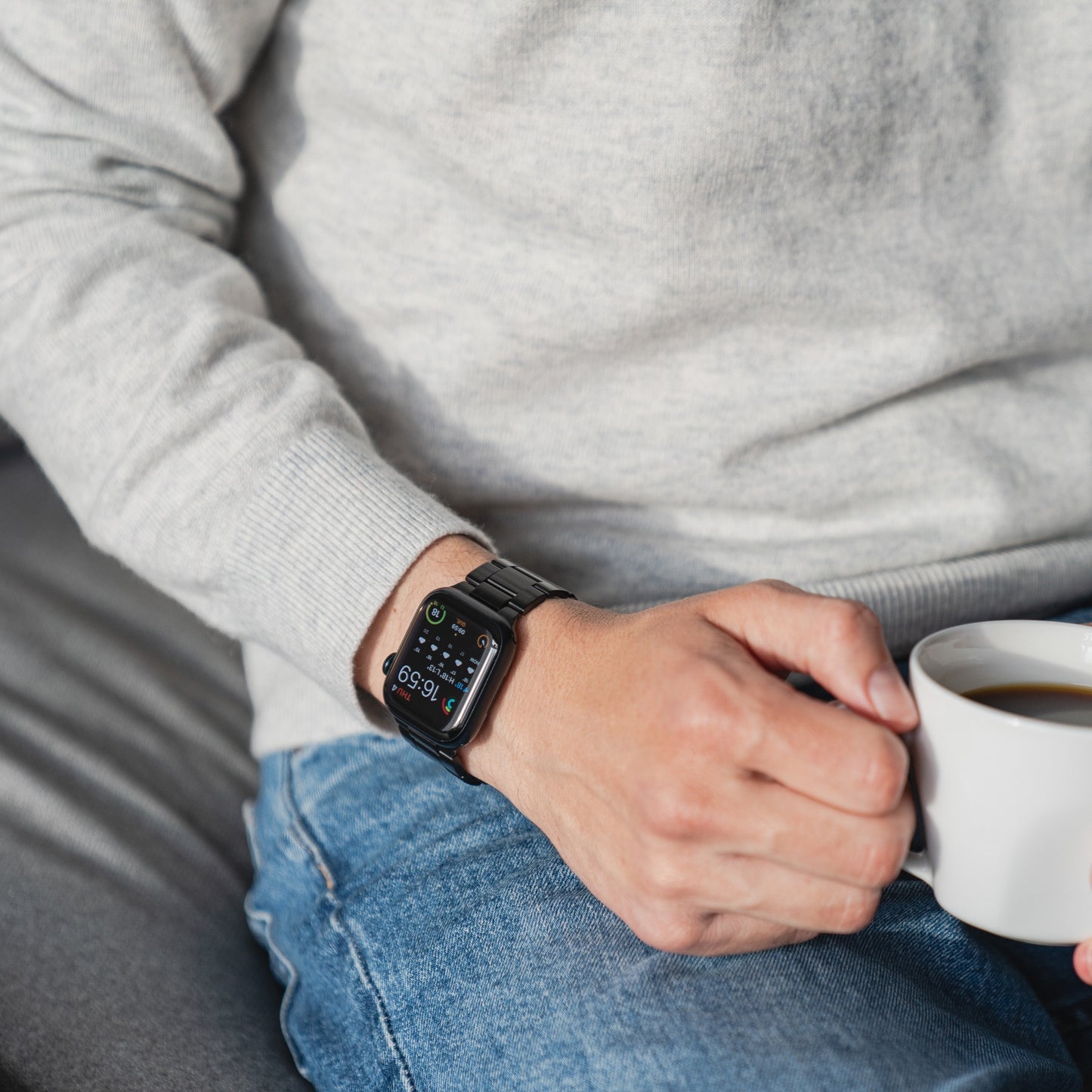 Eine Person in hellgrauem Pullover und Bluejeans sitzt da und hält eine weiße Tasse Kaffee in der Hand. Am linken Handgelenk trägt sie das arktisband Apple Watch Gliederarmband „Prime“ von arktisband, das die Uhrzeit und verschiedene Informationen anzeigt.