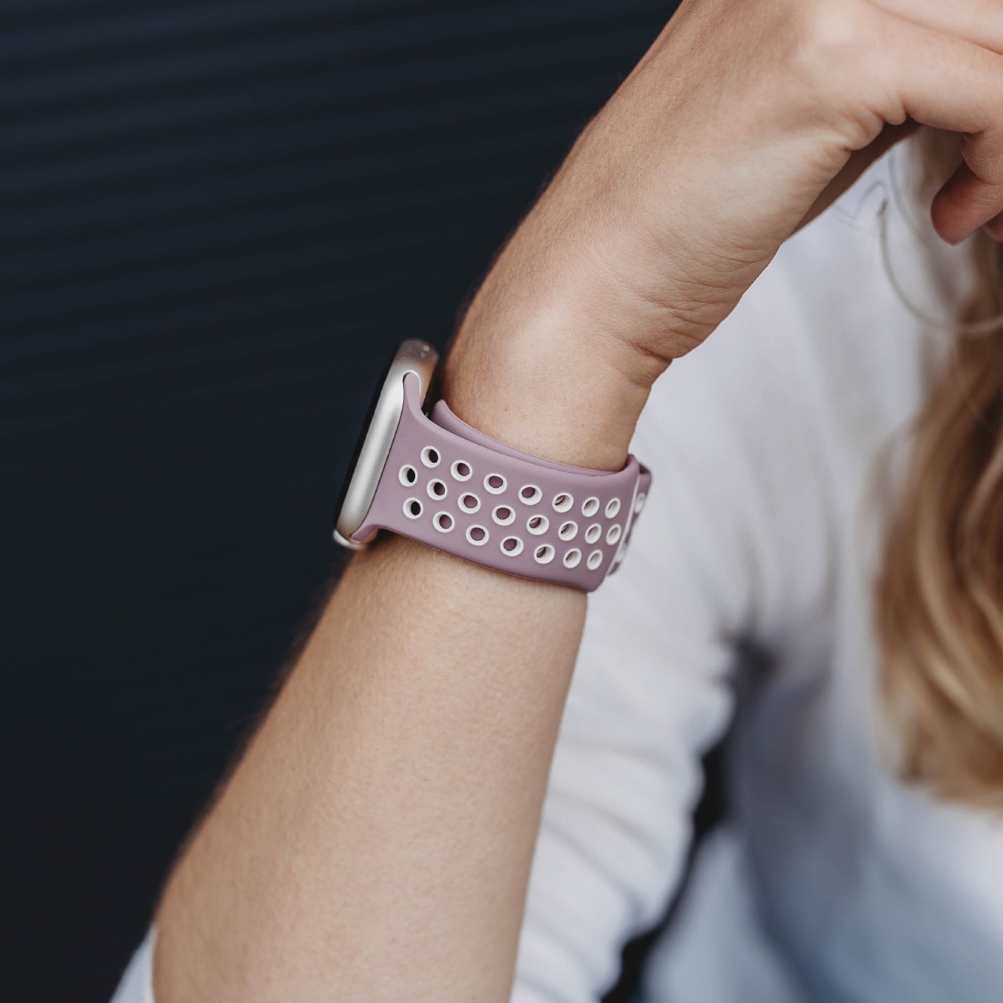 Eine Nahaufnahme zeigt das Handgelenk einer Person, die das arktisband Apple Watch Silikonarmband „Energy“ in Pink und Weiß trägt. Ihr Arm ruht auf einem dunklen, unscharfen Hintergrund, auf dem blonde Haare zu sehen sind.