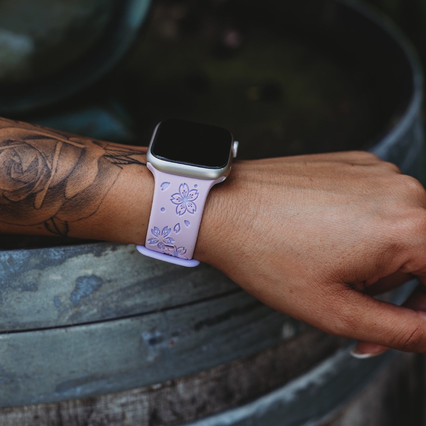 Eine Person mit einem floralen Unterarmtattoo trägt das arktisband Apple Watch Armband „Flores“ in Hellviolett mit floralem Design. Das schweißabsorbierende Band von arktisband liegt auf einer verwitterten, runden Oberfläche und sorgt so für ganztägigen Tragekomfort.