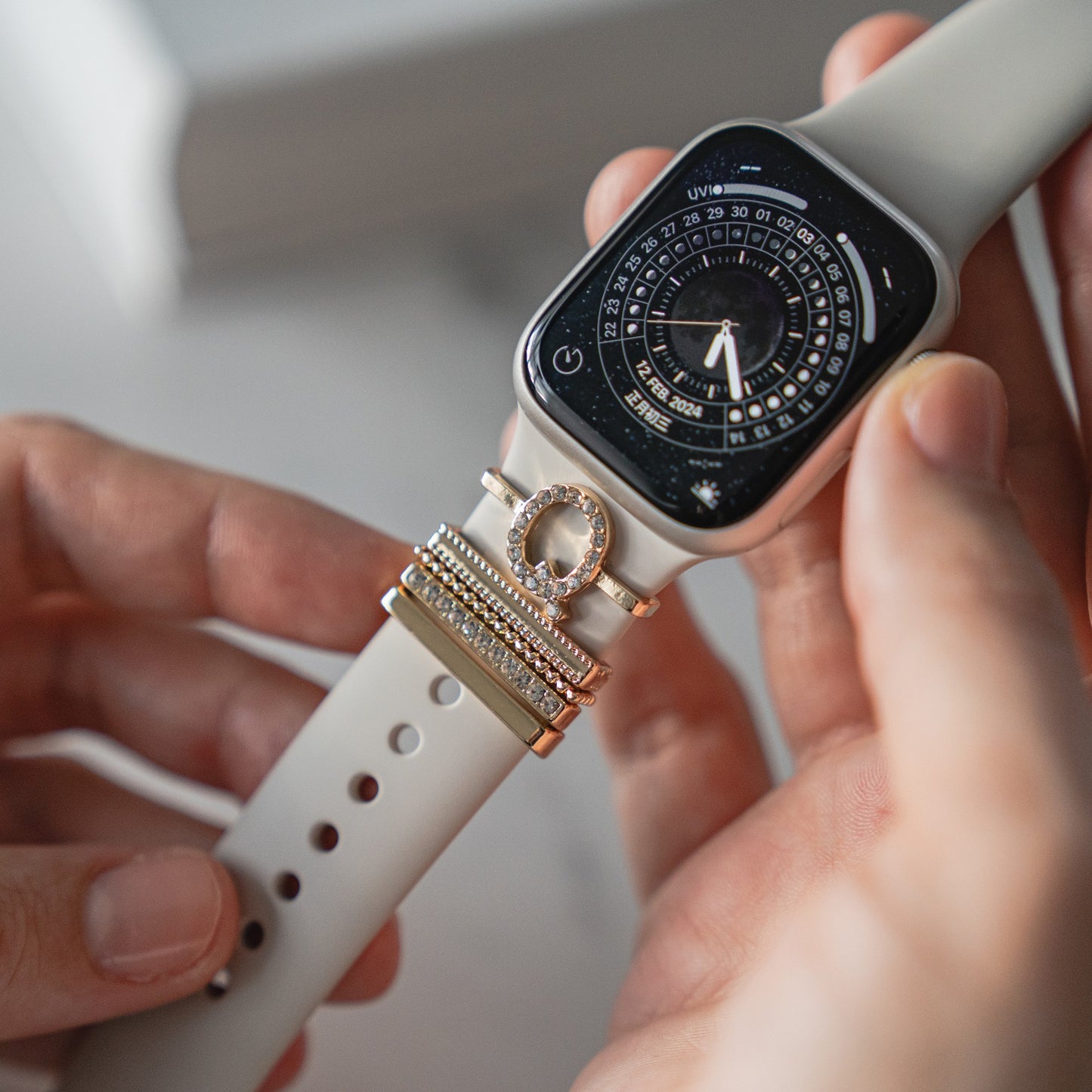 Nahaufnahme von Händen, die eine Apple Watch mit Arktisband Apple Watch Charms „Q Style“ in Gold und Silber auf einem beigen Arktisband-Armband halten.