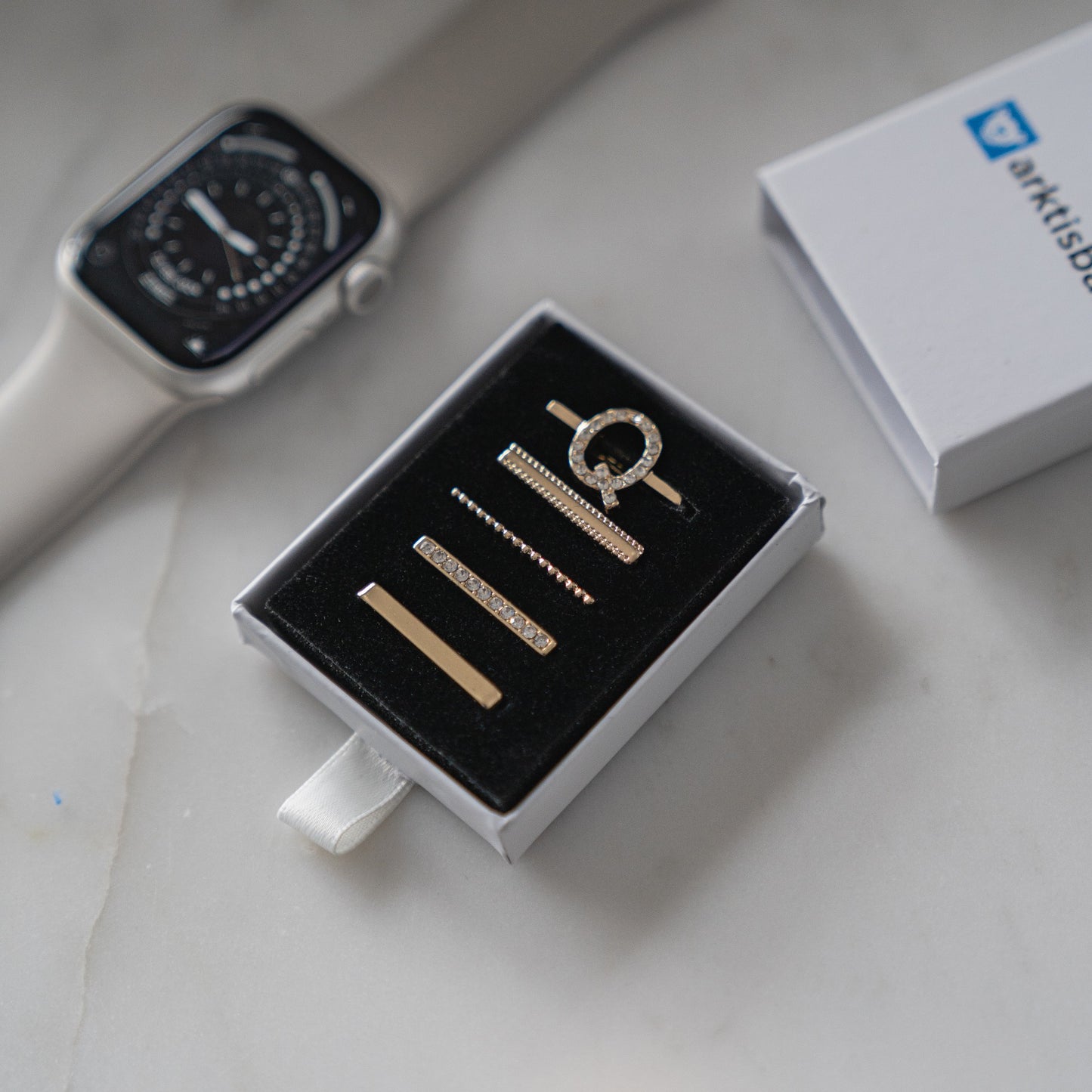 Eine Arktisband Apple Watch und eine Box mit vier metallischen Krawattenklammern, einer eleganten Krawattenklammer und stilvollen Arktisband Apple Watch Charms „Q Style“ liegen auf einer weißen Marmoroberfläche.