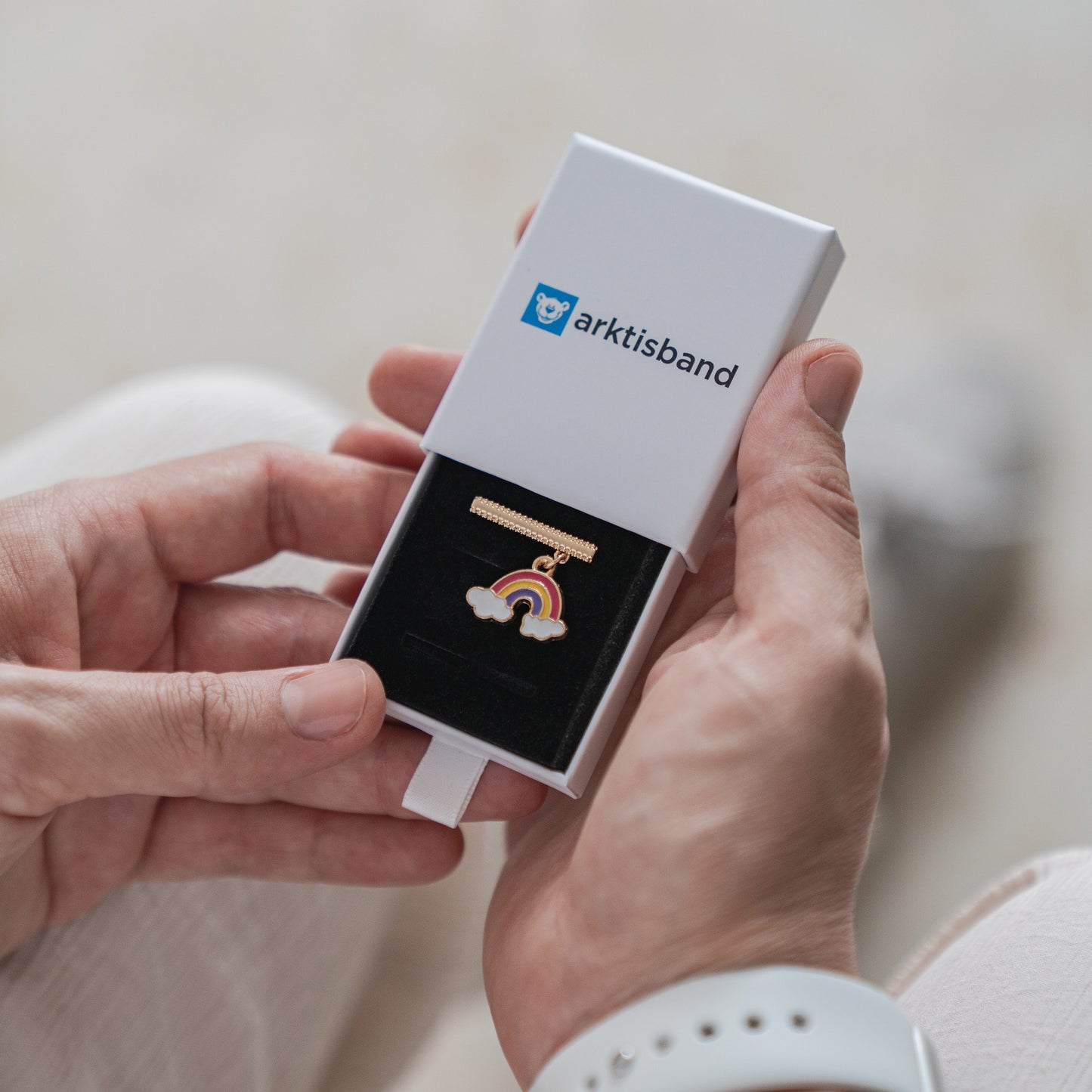 Eine Person hält eine offene weiße Arktisband-Box. Darin befindet sich eine goldene Anstecknadel mit einem Regenbogen- und Wolkenanhänger auf einem schwarzen Kissen – ideal als Ergänzung zu den Arktisband-Armbandanhängern für die Apple Watch. Die Person trägt ein weißes Armband.