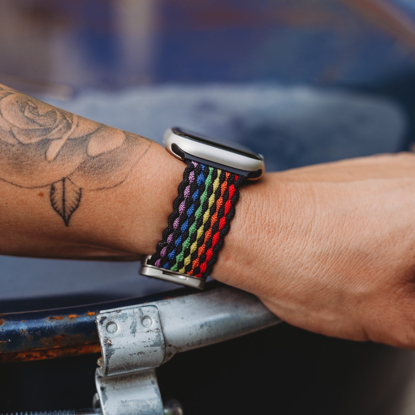 Eine Person mit einem floralen Unterarmtattoo trägt das arktisband Geflochtene Apple Watch Armband „Casual“ Rainbow Edition, ihre Hand ruht auf einer Metalloberfläche.