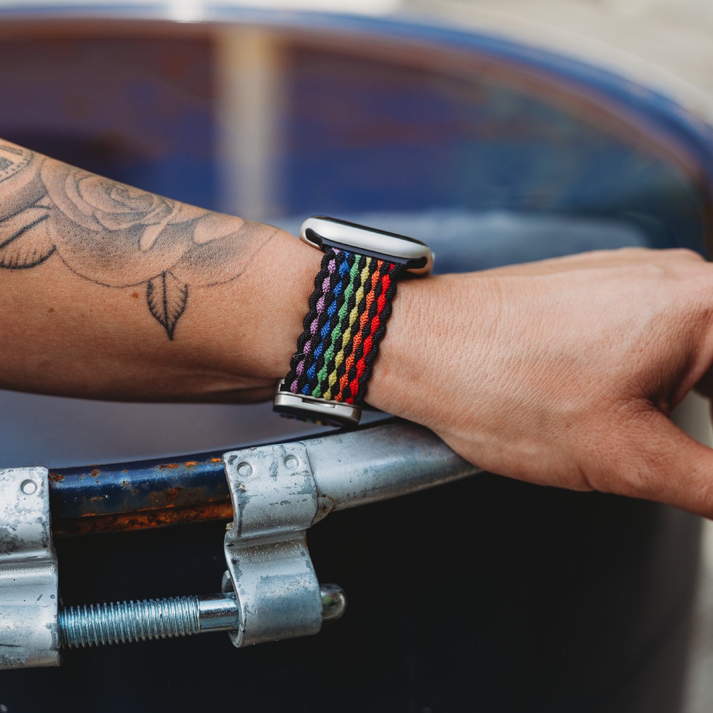 Eine Nahaufnahme eines Handgelenks, das das geflochtene Apple Watch Armband „Casual“ Rainbow Edition von arktisband trägt. Die Person mit einem Rosentattoo auf dem Unterarm greift eine Metallstange mit abgeplatzter blauer Farbe.