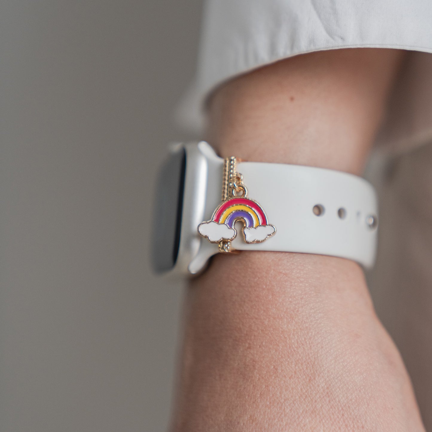 Eine Nahaufnahme eines Handgelenks mit einem Arktisband-Apple-Watch-Armband mit einem Arktisband-Anhänger mit einem kleinen Regenbogen und einer Wolke, kombiniert mit einem weißen Langarmshirt.