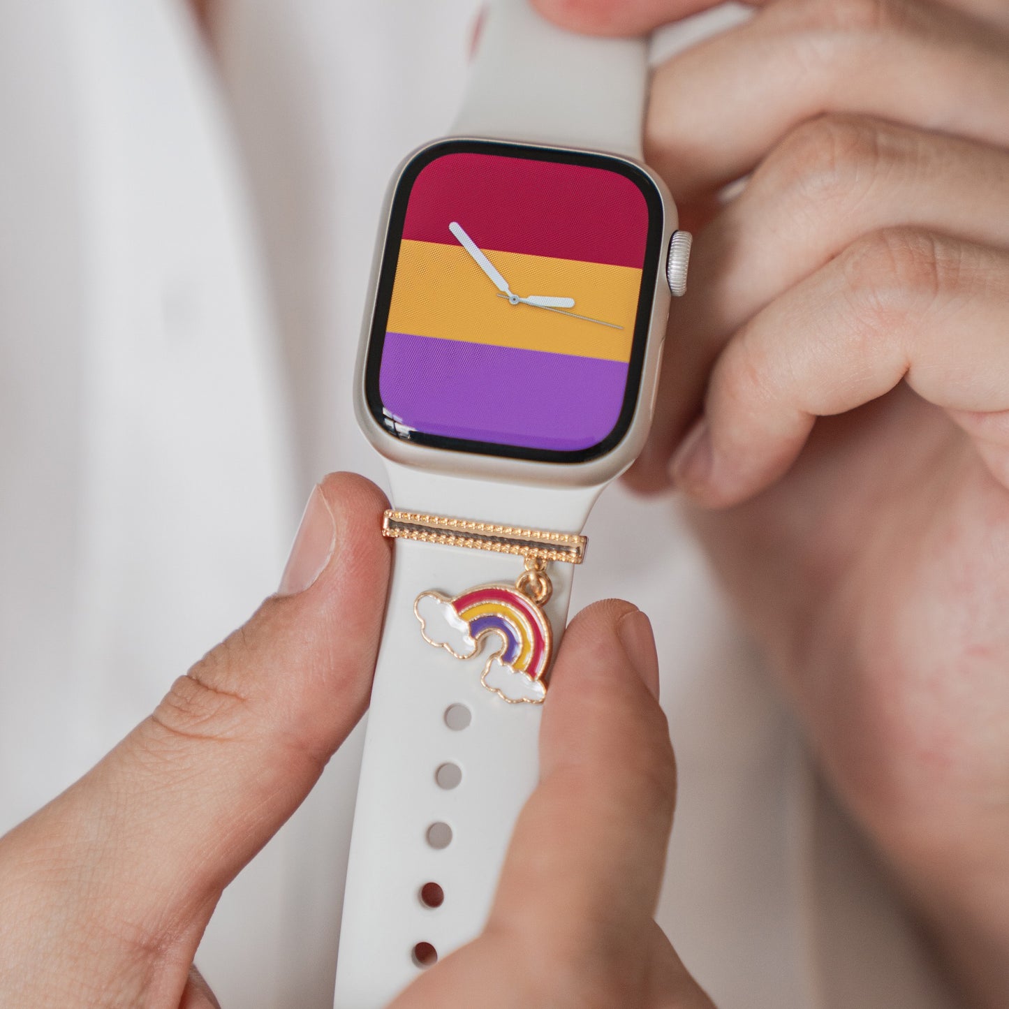 Eine Person passt eine weiße Apple Watch an, deren Display eine Flagge mit violetten, gelben und roten Streifen zeigt. Das Armband ist mit den arktisband Apple Watch Armband Charms – einem Regenbogen und einer Wolke – für einen einzigartigen Look personalisiert.