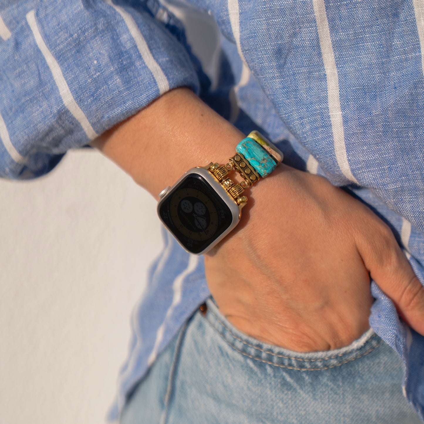 Person trägt eine Arktisband Apple Watch „Stone Loop“ von Arktisband und bunte Perlenarmbänder, die Hand in der Tasche, bekleidet mit einem blau gestreiften Hemd und Jeans.