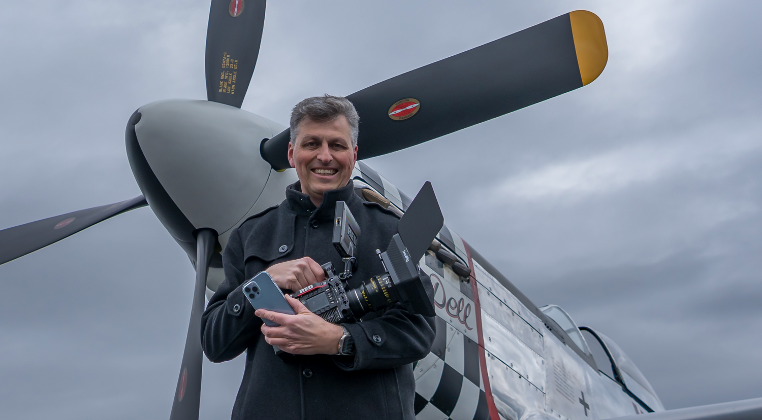 Rainer Wolf steht mit einer RED Filmkamera und einem iPhone in der Hand vor einem Mustang P 52  Flugzeug