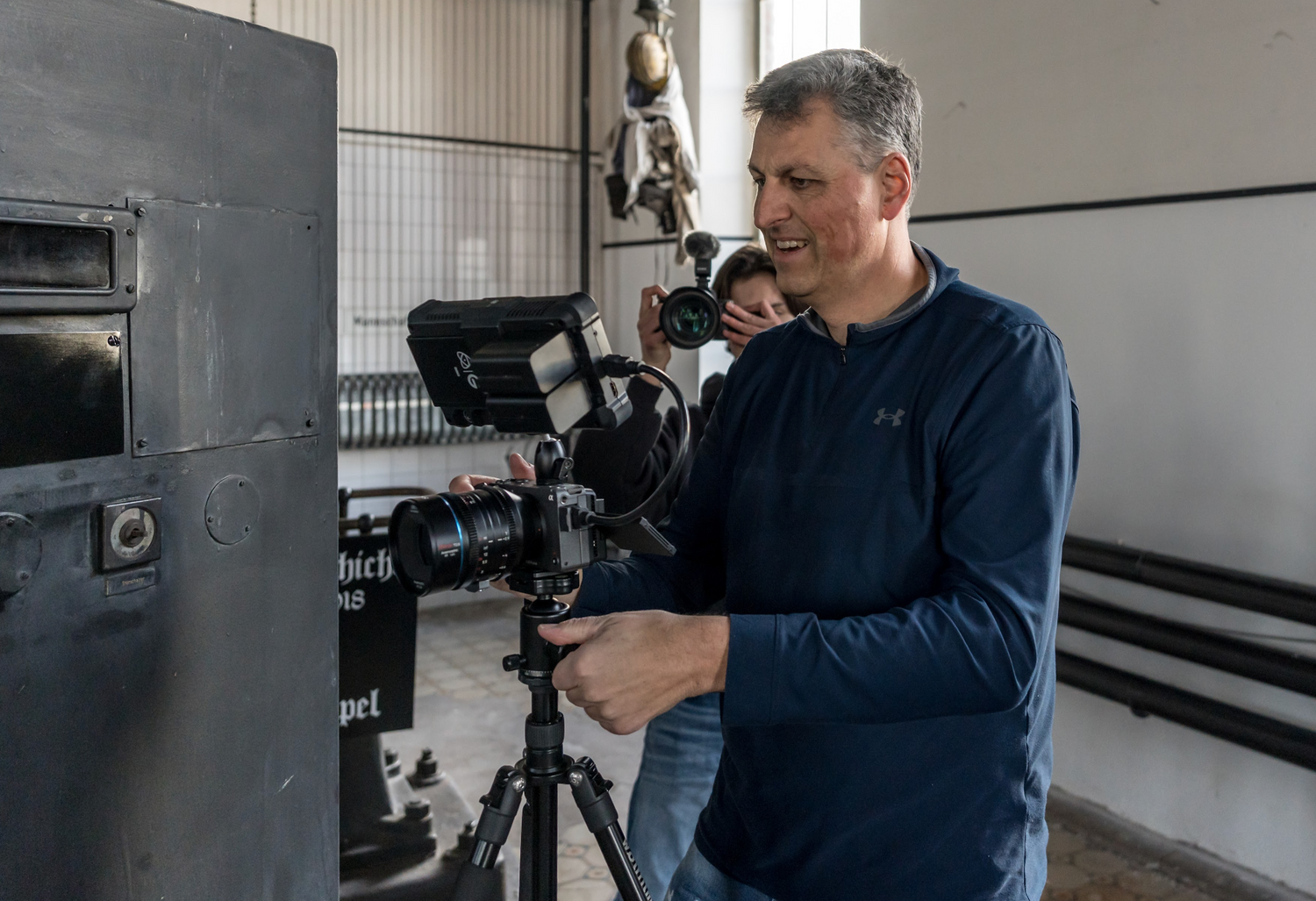 Mann filmt in einem Gebäude mit einer professionellen Kamera auf einem Stativ; eine andere Person hält eine Kamera hinter ihm.