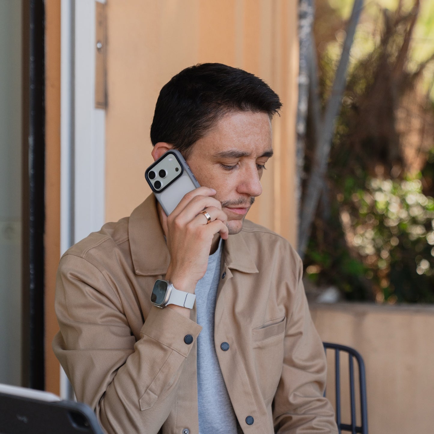 Ein Mann mit hellbrauner Jacke sitzt im Freien und spricht mit seinem Smartphone, geschützt durch das Arktis iPhone 17 Pro Solid Case, eine matte, stoßabsorbierende Hülle von Arktis.