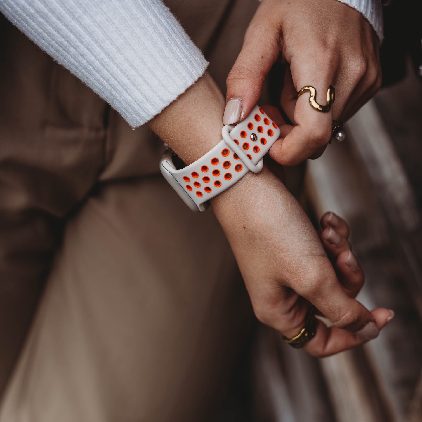 Eine Person mit manikürten Nägeln und goldenen Ringen rückt ein Arktisband Apple Watch Silikonarmband „Energy“ in Weiß mit orangefarbenen Löchern an ihrem linken Handgelenk zurecht, während sie ein weißes Langarmshirt trägt.