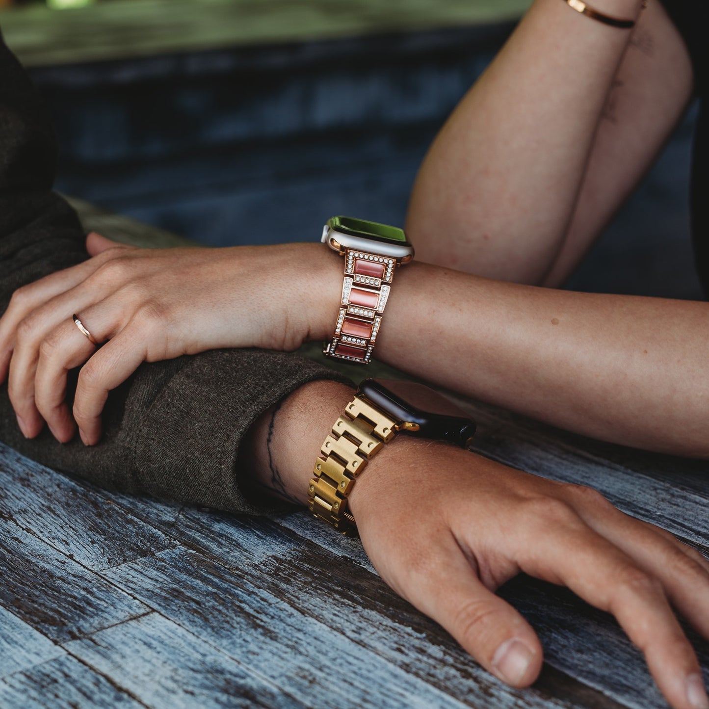 Zwei Personen stützen ihre Arme auf einer Holzfläche ab. Die eine trägt das arktisband Apple Watch Gliederarmband „Senorita“, ein elegantes pink-goldenes Edelstahlarmband, die andere eine klassische goldene Uhr mit schmalem Ring.