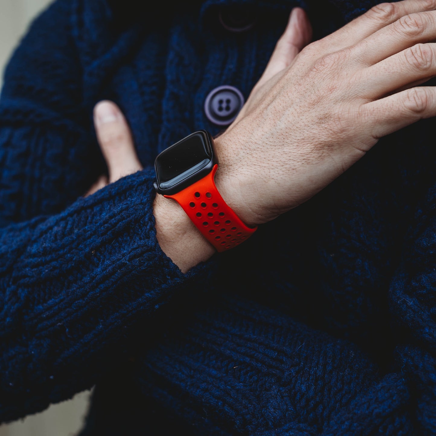 Eine Person in einem dunkelblauen Strickpullover verschränkt die Arme und präsentiert das arktisband Apple Watch Silikonarmband „Energy“ von arktisband mit einem rot-schwarzen Lochmuster am Handgelenk.
