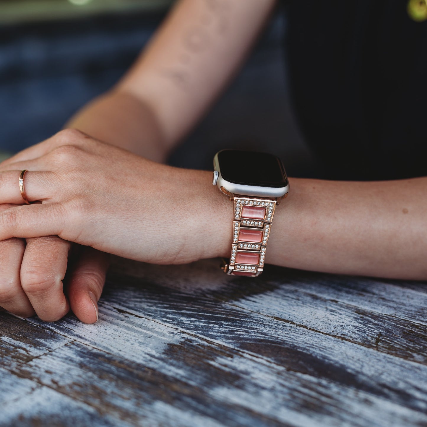 Eine Person mit einem goldenen Ring legt ihre Hände auf einen Holztisch und trägt das arktisband Apple Watch Gliederarmband „Senorita“, ein roségoldenes, mit Strasssteinen besetztes Metallarmband von arktisband.