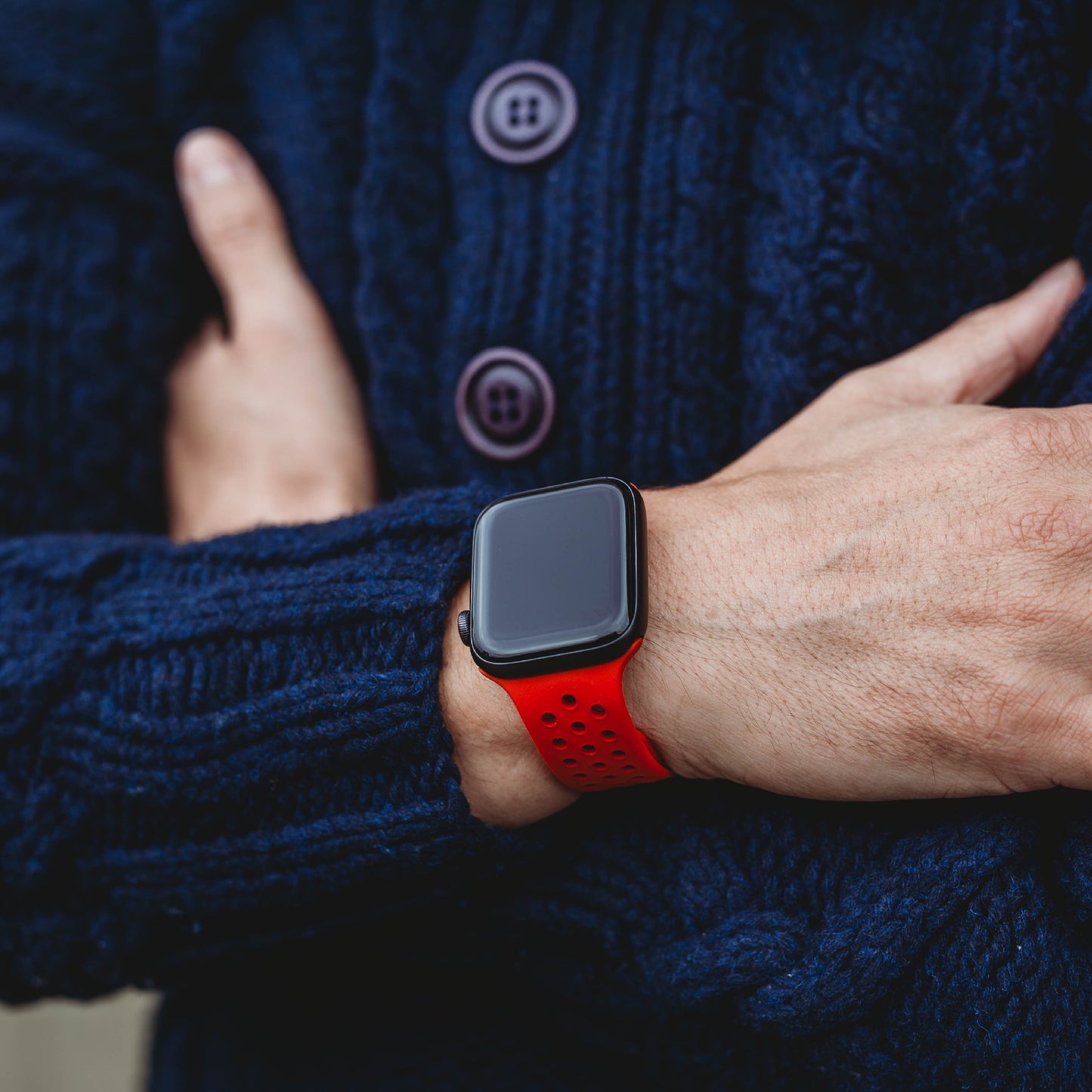 Eine Person in einem dunkelblauen strukturierten Pullover mit großen Knöpfen verschränkt die Arme und präsentiert das Arktisband Apple Watch Silikonarmband „Energy“ an ihrem Handgelenk, mit einem roten perforierten Band und einem schwarzen Bildschirm.