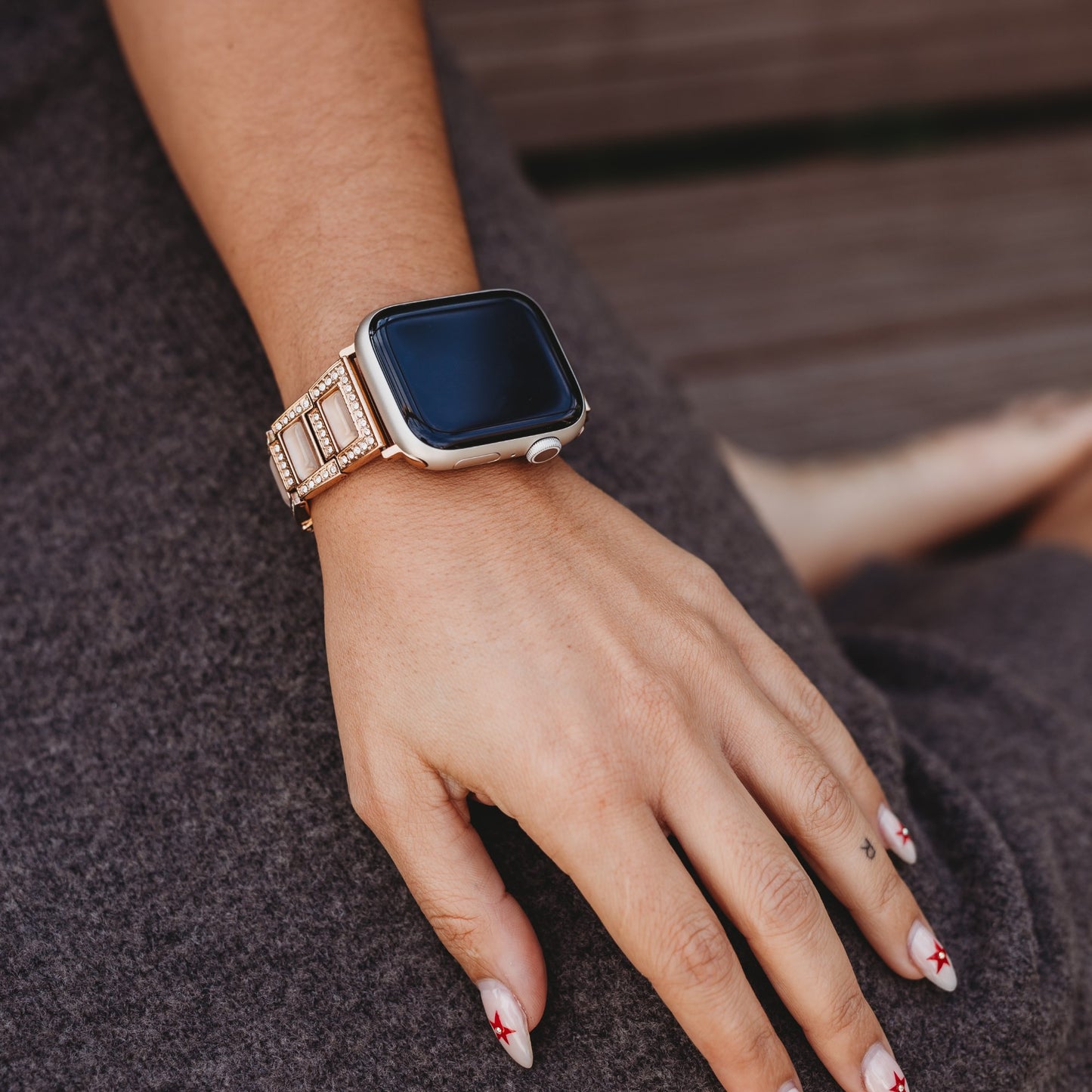 Eine Person, die ein arktisband Apple Watch Gliederarmband „Senorita“ von arktisband trägt, legt ihre Hand auf den Schoß; hautfarbene Nägel mit roten Kreuzen heben sich vom grauen Stoff und dem Holzhintergrund ab.