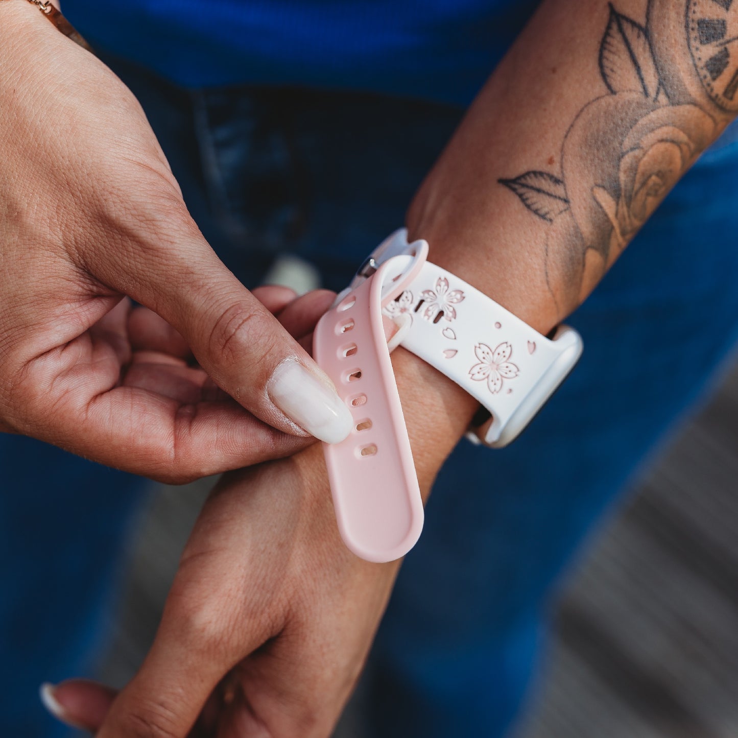 Eine Person in Blue Jeans und mit einem Blumentattoo auf dem Unterarm befestigt das rosa-weiße arktisband Apple Watch Armband „Flores“ von arktisband an ihrer Apple Watch.