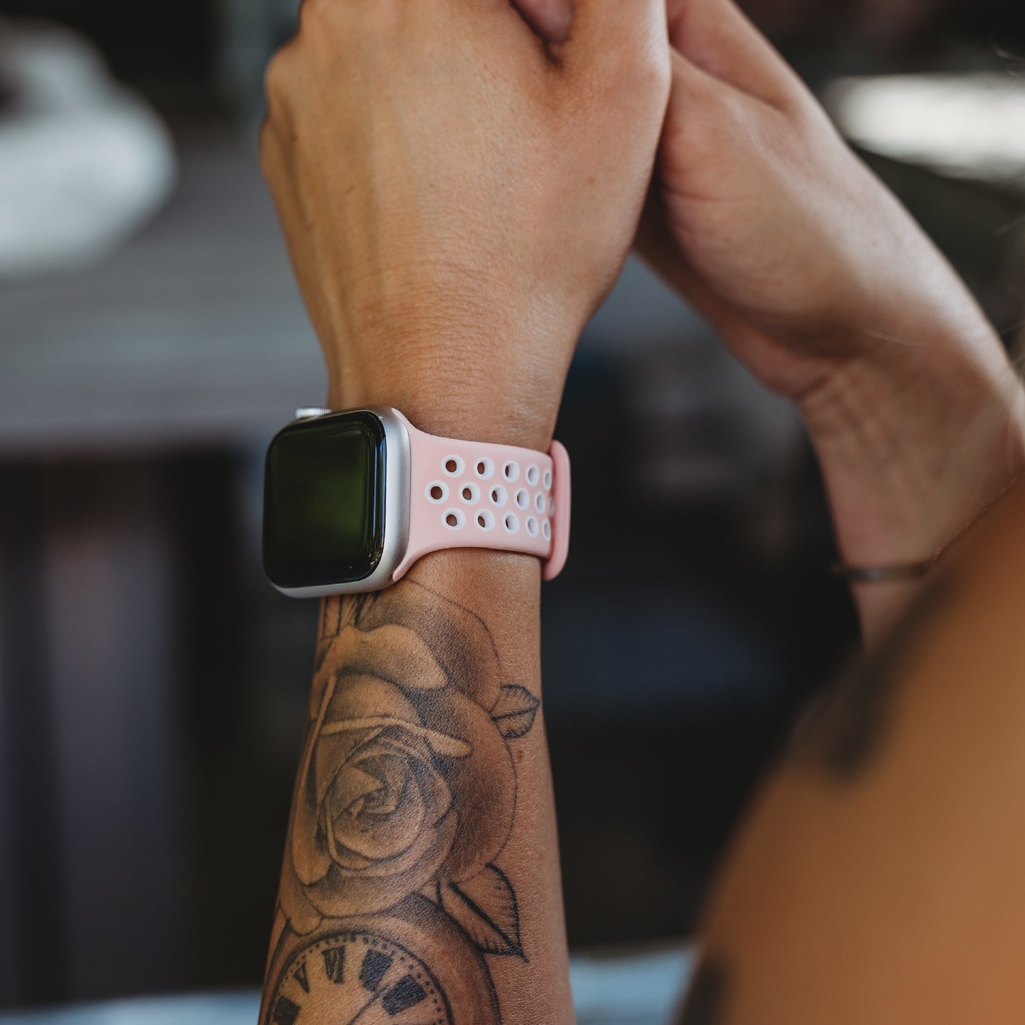 Jemand trägt ein arktisband Apple Watch Silikonarmband „Energy“ in Pink und Weiß von arktisband am Handgelenk, gepaart mit einem Rosen- und Uhrentattoo auf dem Unterarm, während im Hintergrund die Hände zusammengehalten werden.