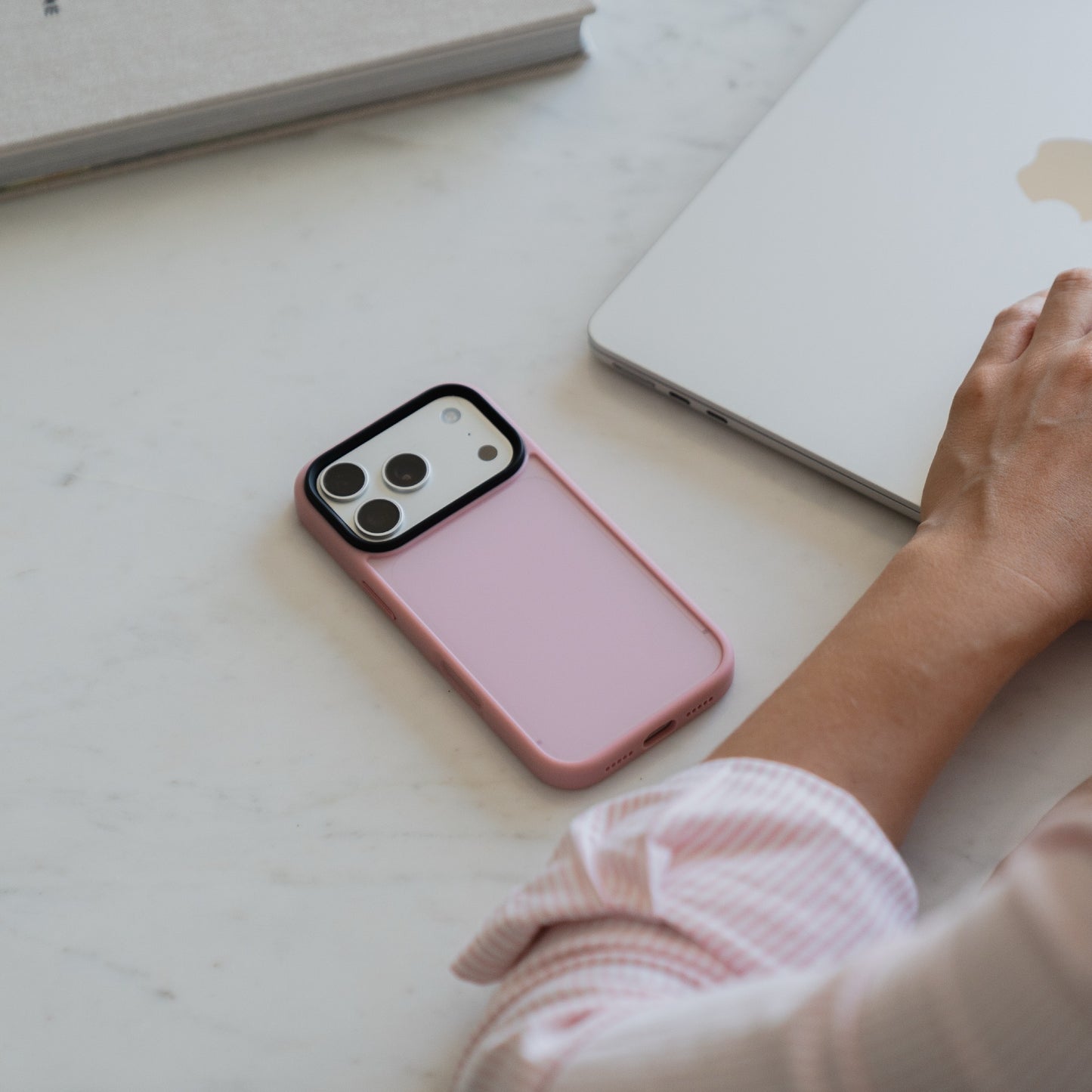 Ein Arktis iPhone 17 Pro Solid Case in Pink mit mattem Finish liegt neben der Hand einer Person, die einen Laptop auf einem weißen Schreibtisch benutzt.