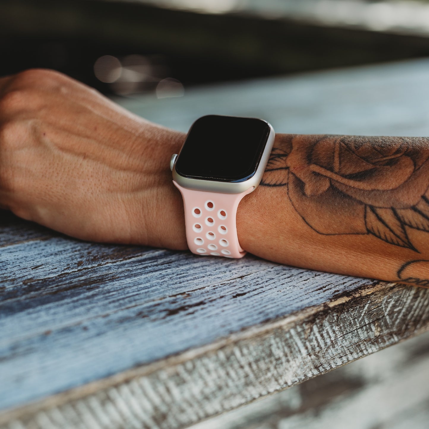 Eine Person mit einem Blumentattoo auf dem Unterarm trägt ein Arktisband Apple Watch Silikonarmband „Energy“ in Hellrosa mit Löchern, während ihr Arm auf einer verwitterten Holzoberfläche ruht.