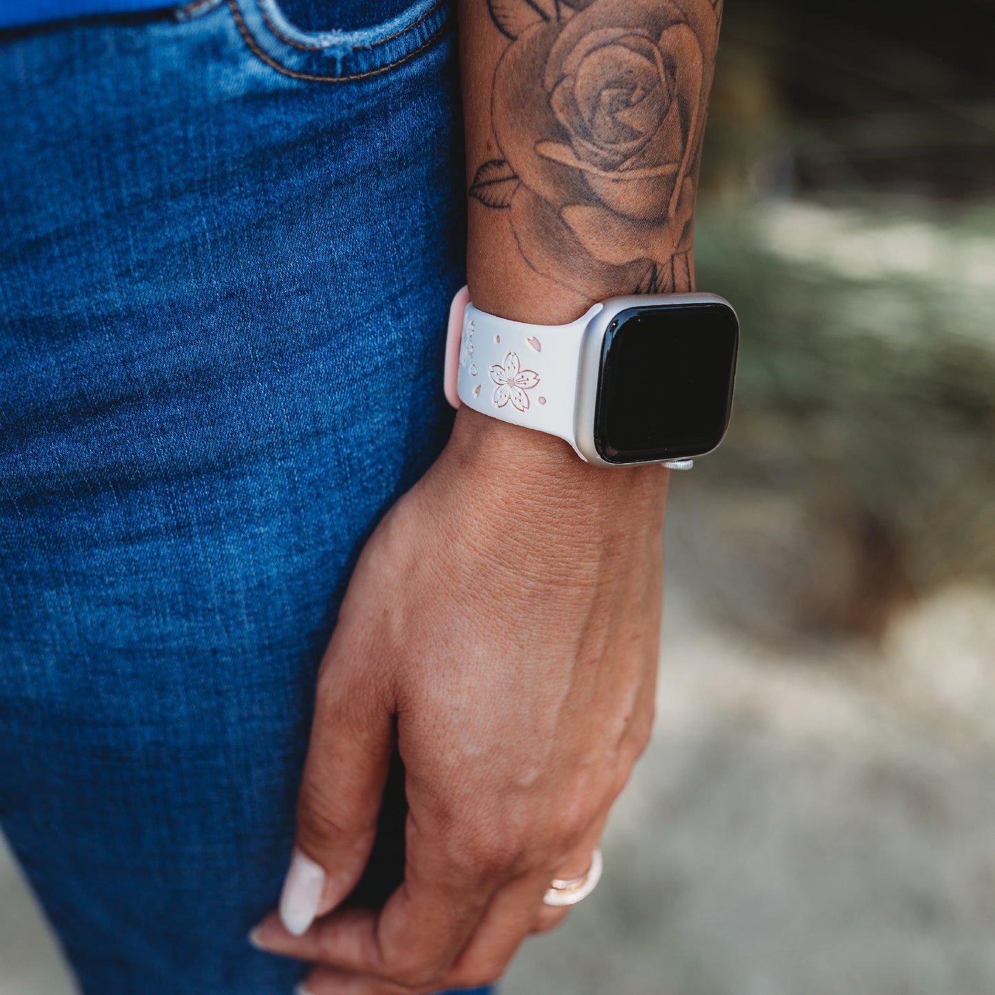 Eine Person in Blue Jeans trägt ein arktisband Apple Watch Armband „Flores“ von arktisband. Ihr mit einer Rose tätowierter Arm hängt entspannt an ihrer Seite.