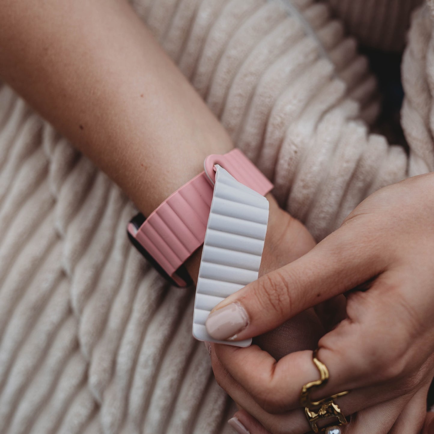 Eine Person trägt das arktisband Apple Watch Magnetarmband „Neo“ in Pink mit weißem, geripptem Aufsatz am Handgelenk. Der goldene Schmuck und der cremefarbene Stoff betonen das stilvolle Magnetarmband. Kompatibel mit Apple Watch.