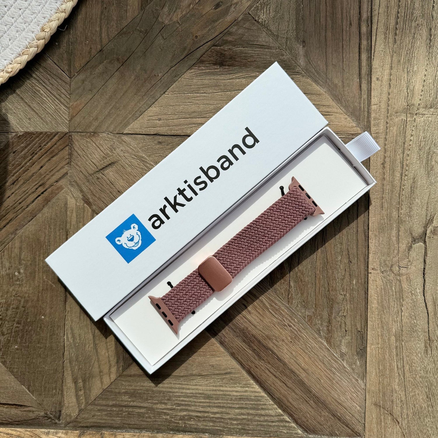 Ein arktisband Geflochtenes Apple Watch Armband „Casual“ mit Magnetverschluss in Braun liegt in einer offenen weißen arktisband Box auf einer Holzoberfläche.