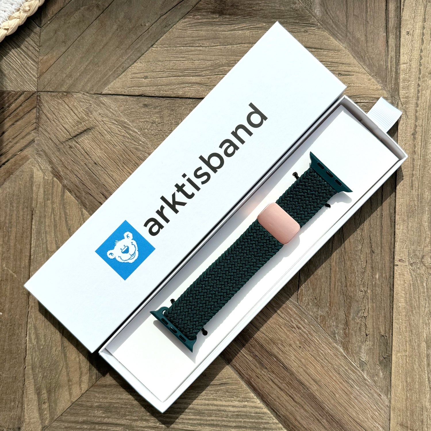 Ein dunkelgrünes arktisband Geflochtenes Apple Watch Armband „Casual“ mit Magnetverschluss ruht in einer offenen weißen arktisband Box auf einer Holzoberfläche.